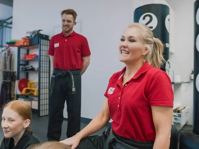 A smiling woman with glasses, hoop earrings, and layered necklaces in an indoor setting.