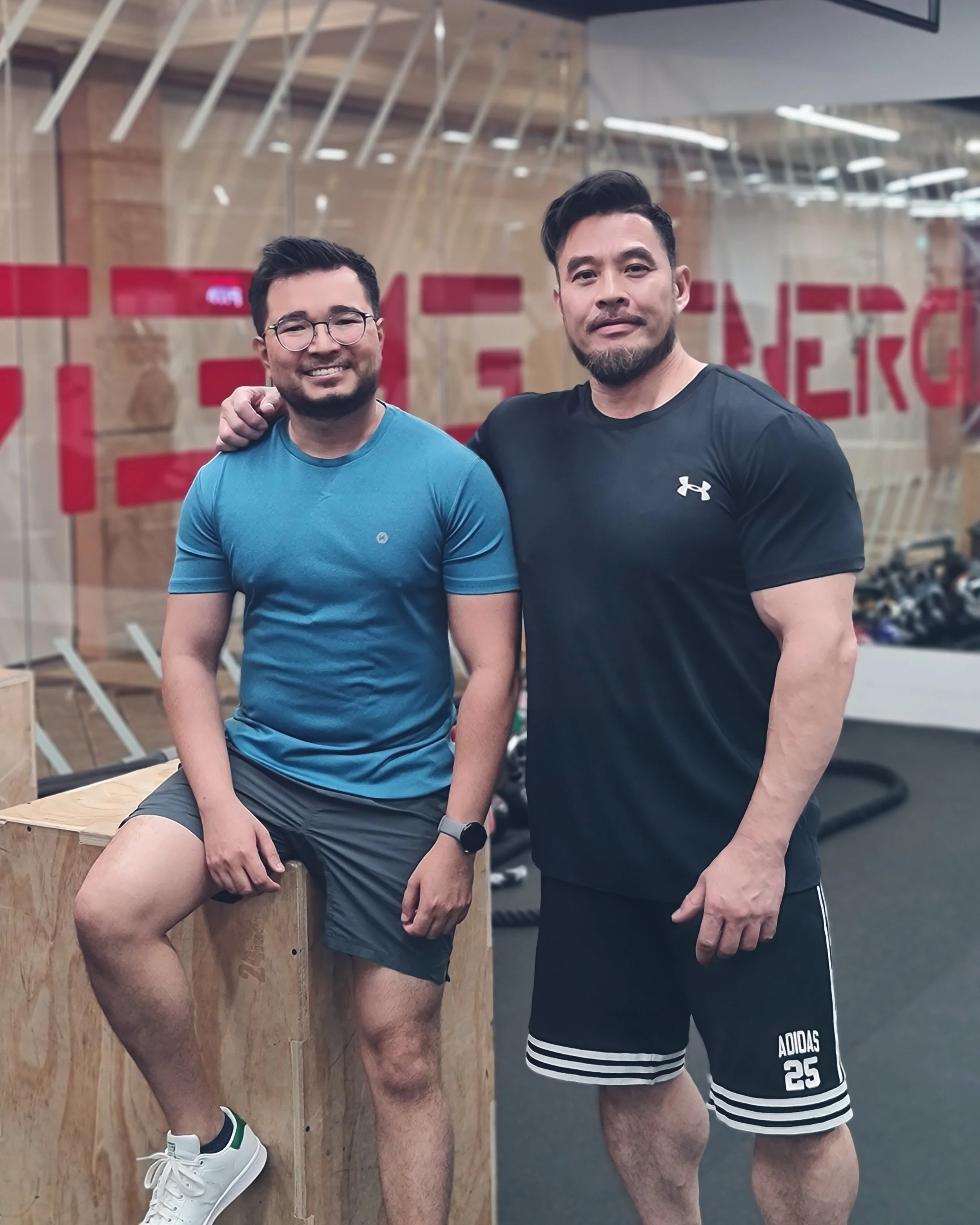 Two men in workout clothes posing inside a gym, one smiling and wearing glasses, the other with a serious expression, with gym equipment in the background.
