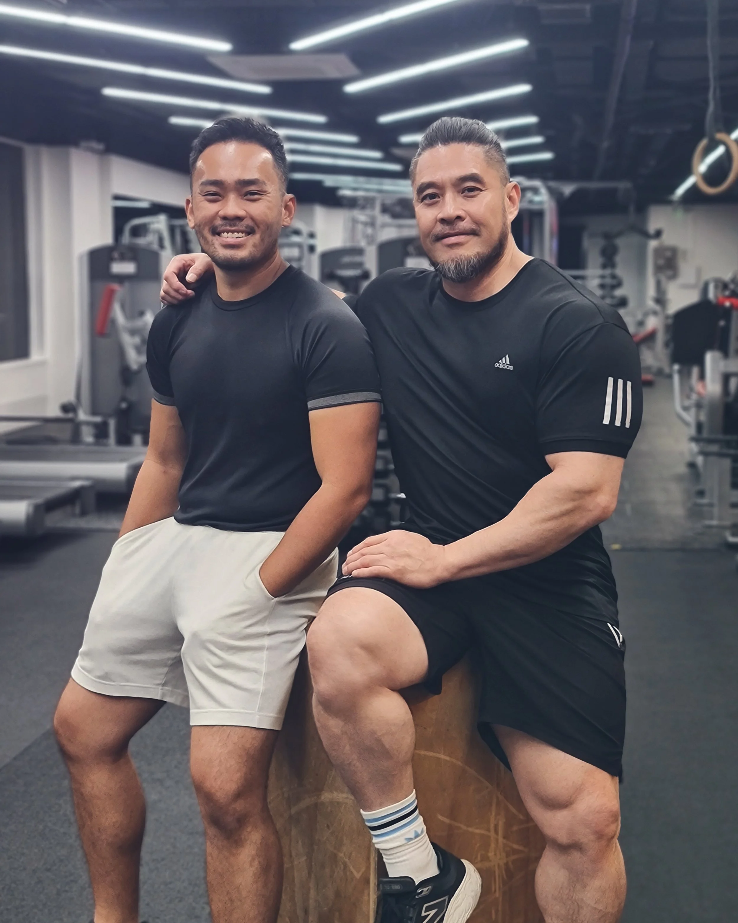 Two men in black athletic shirts posing together in a gym, one sitting on a wooden box with his leg raised, the other standing with his arm around the other man's shoulder.