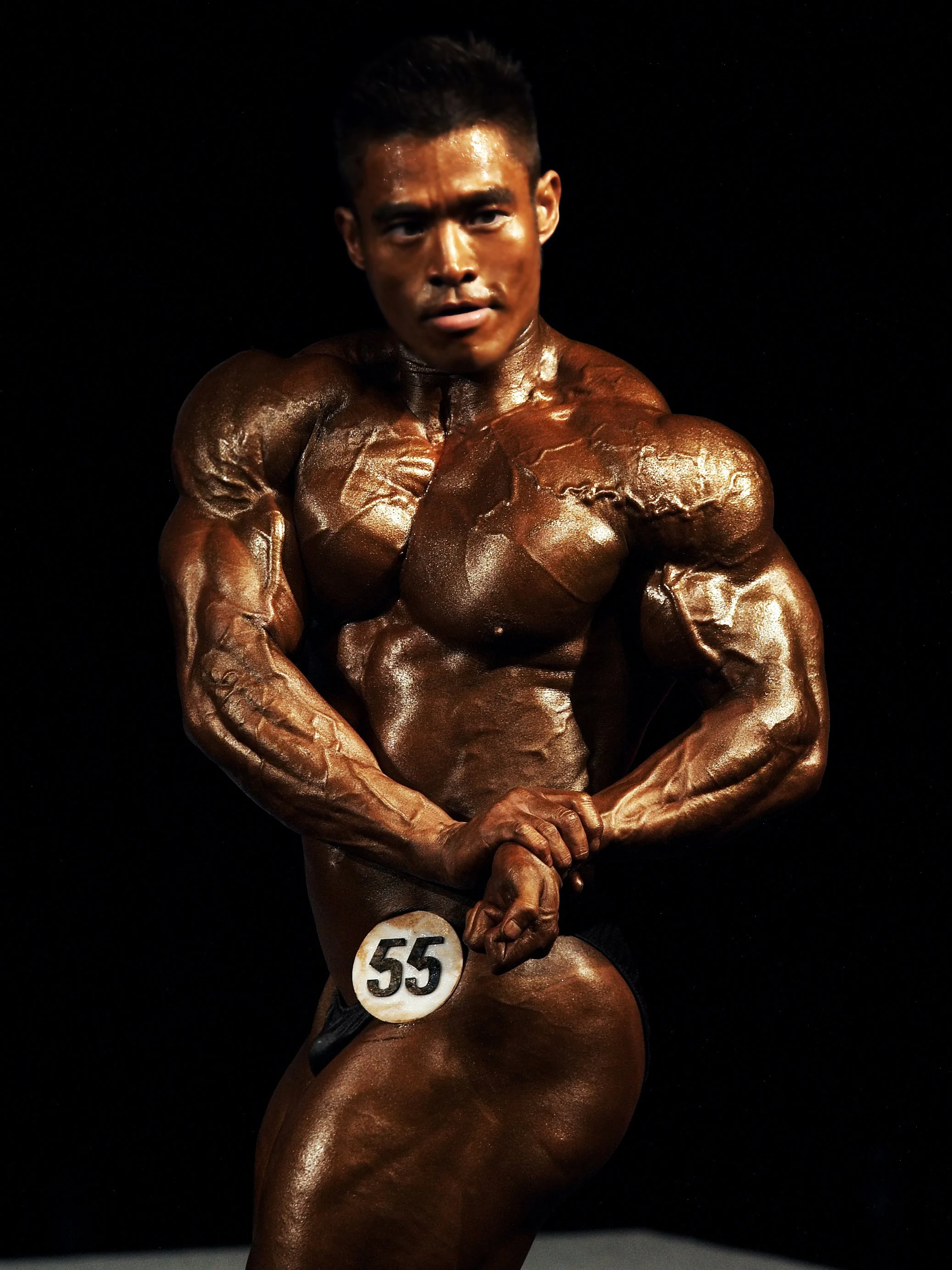 A male bodybuilder with dark hair and tanned, muscular physique, wearing a small black bikini with the number 55, posing on stage against a black background.