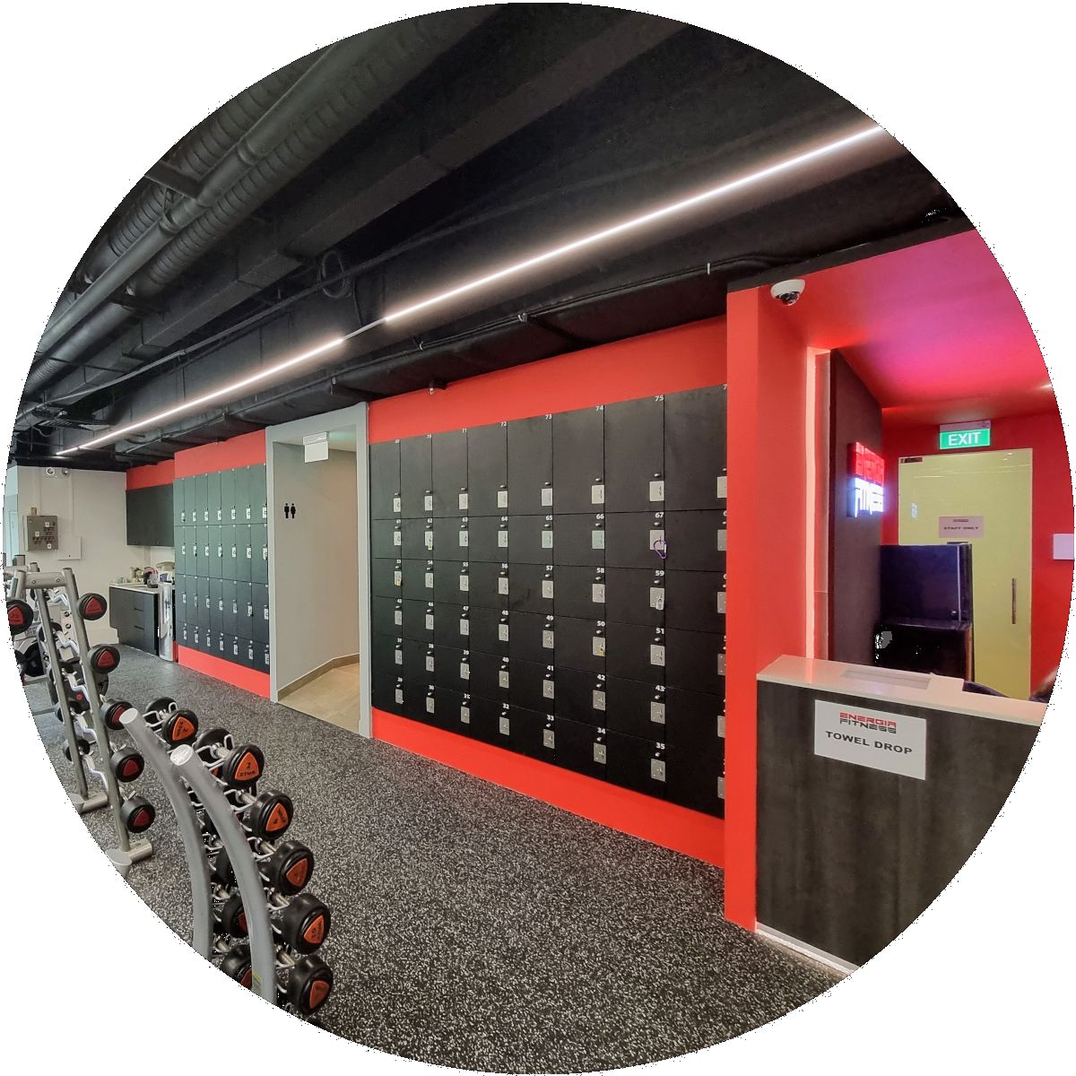 Locker room with black lockers on a red accent wall, a row of dumbbells on a rack, and a towel drop station near an exit door.