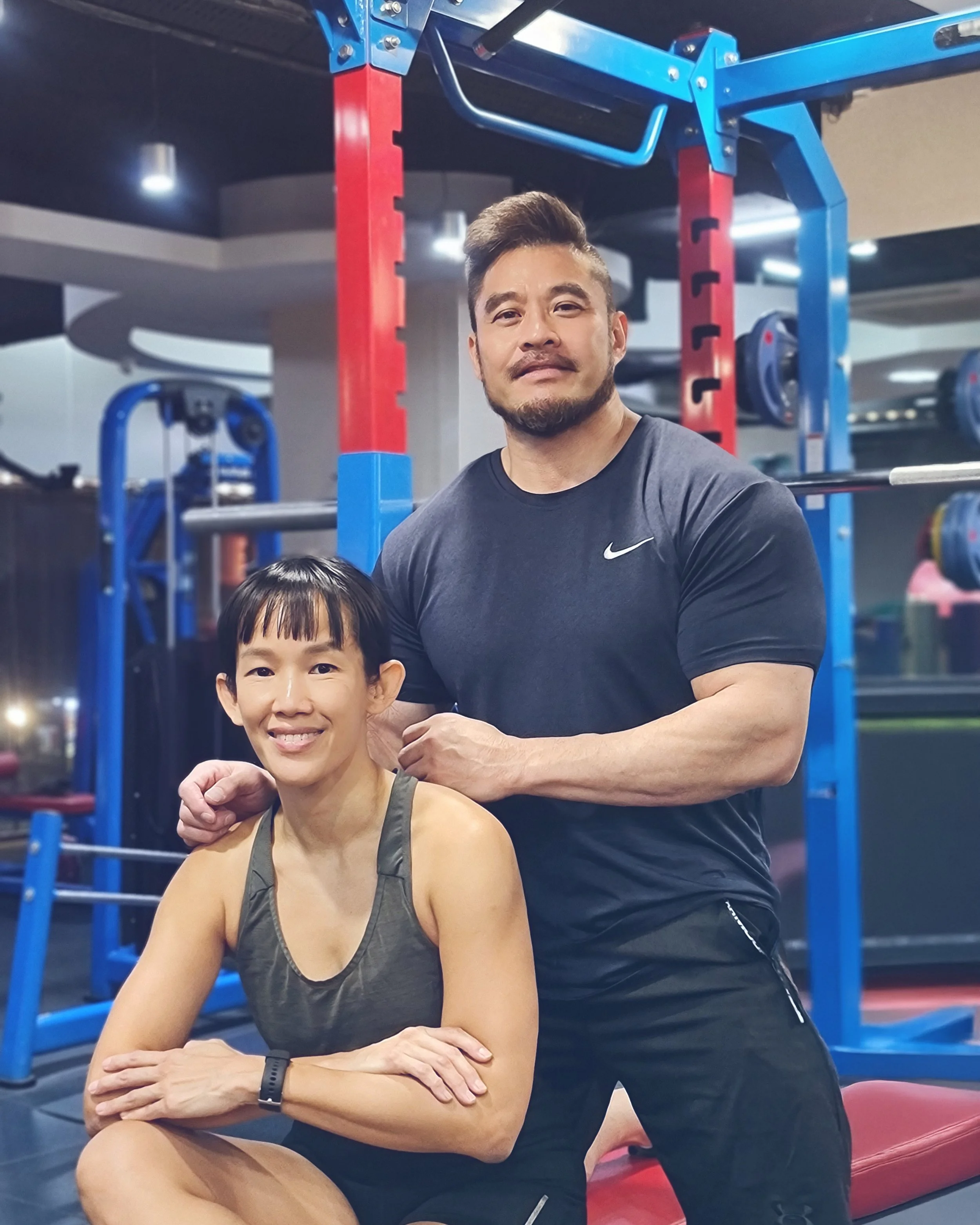 A man standing behind a woman sitting on the floor in a gym, both smiling. The man is wearing a black Nike t-shirt and the woman is wearing a dark athletic tank top.