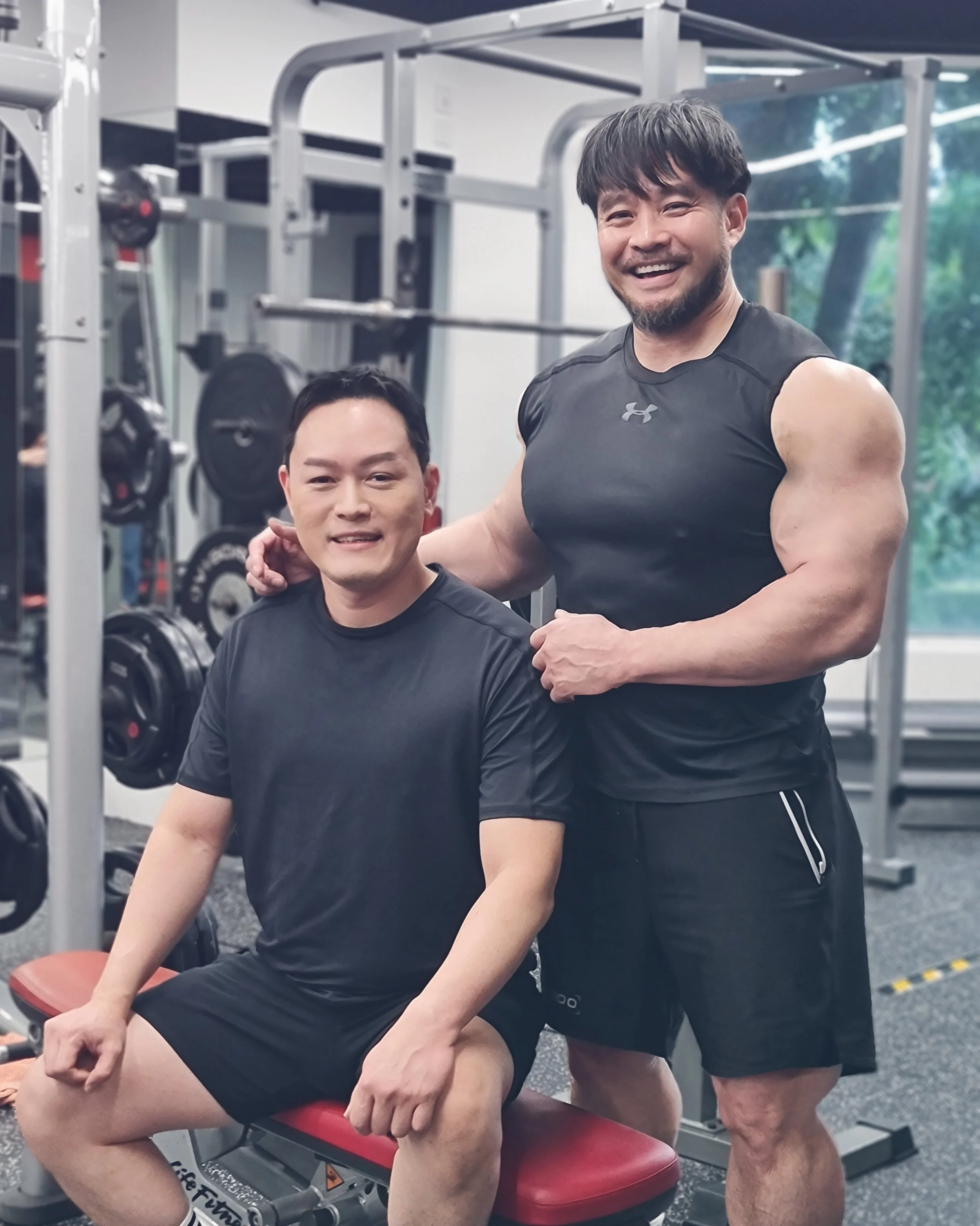 Two men in a gym, one sitting on a workout bench and the other standing behind him with his hand on his shoulder, smiling for the camera.