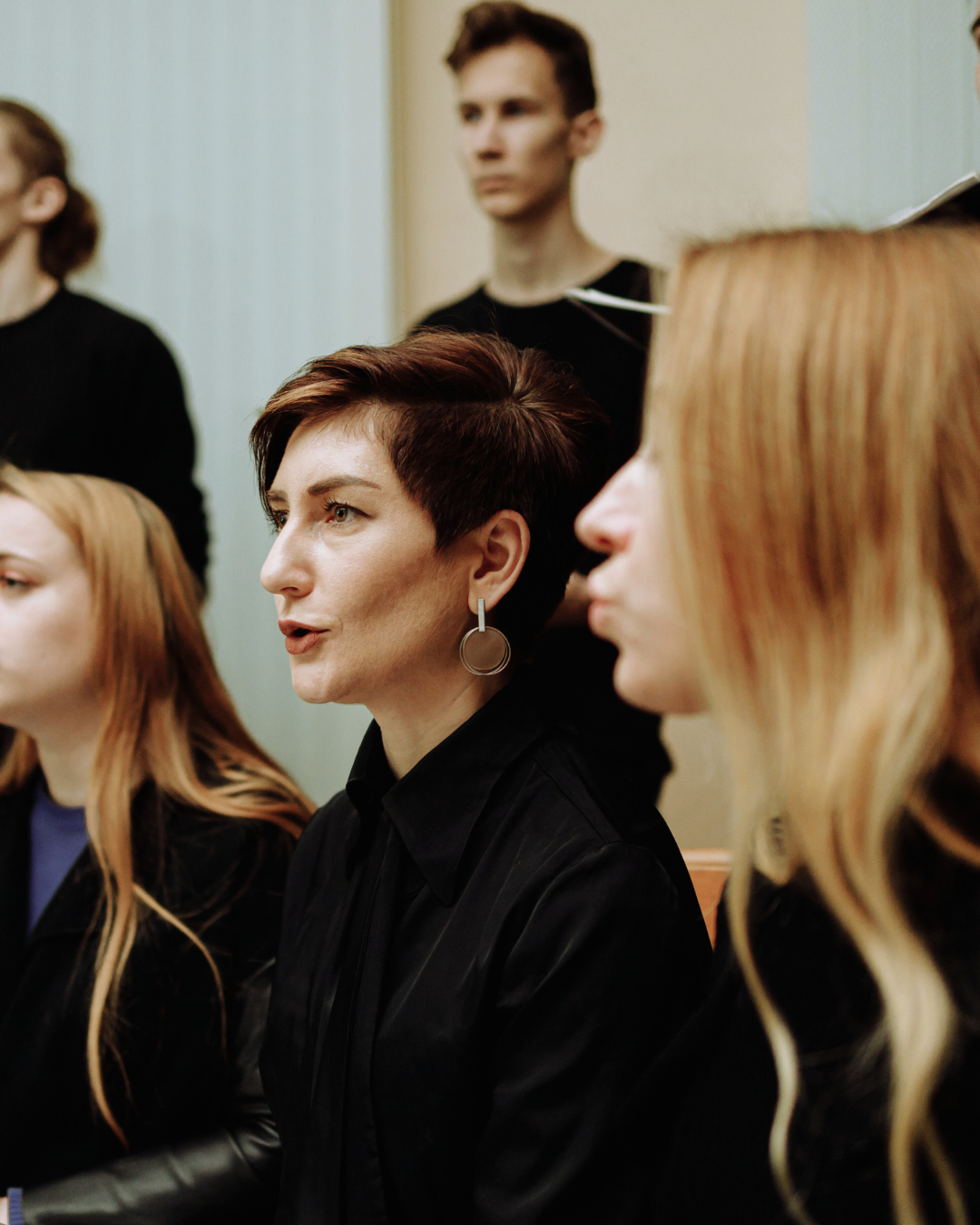Group of women, some with red hair, sitting and listening to a woman with short dark hair speaking, in a room with neutral-colored walls.