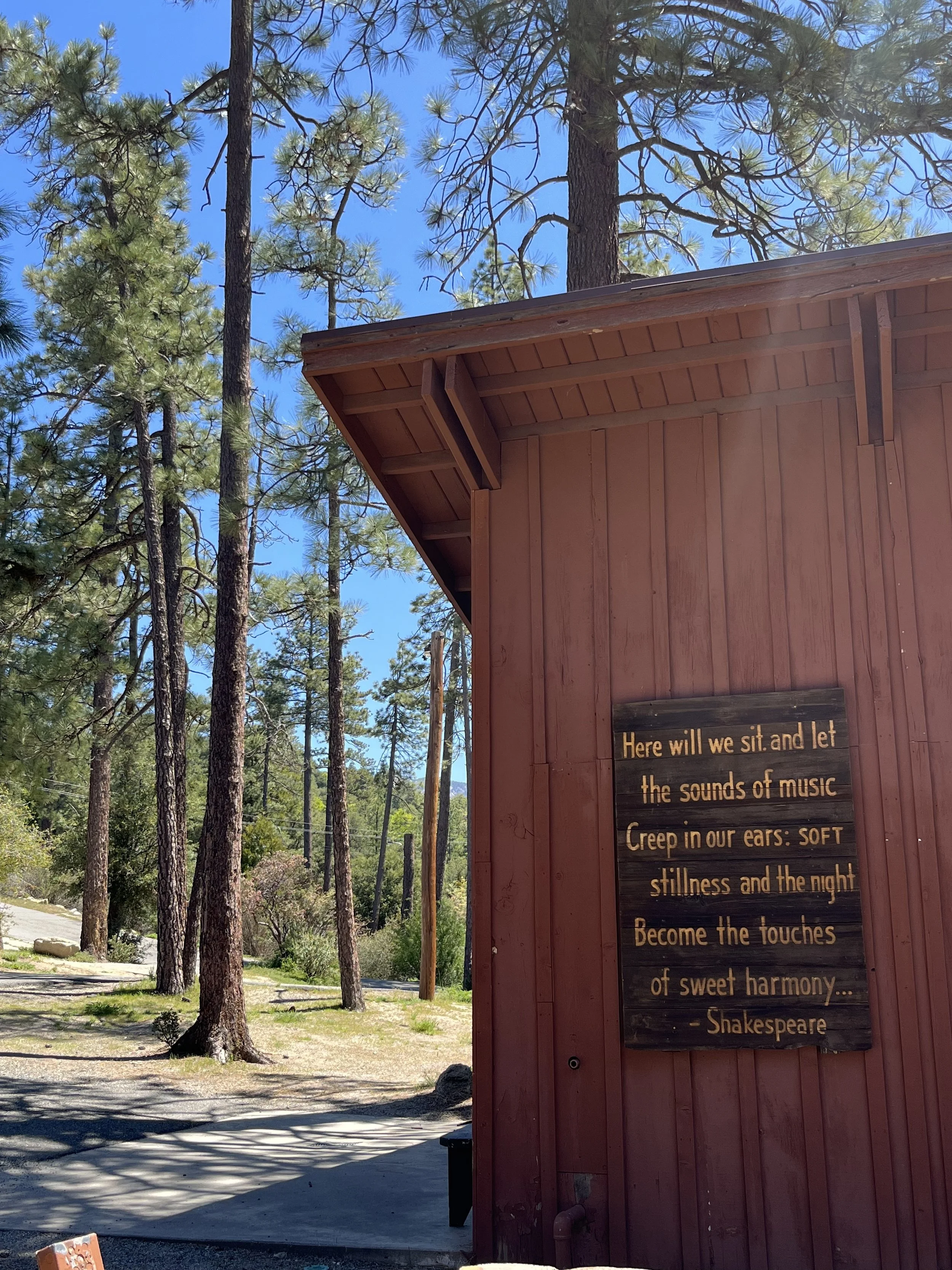 A wooden building with a sign featuring a Shakespeare quote about music and harmony, surrounded by tall pine trees under a clear blue sky.