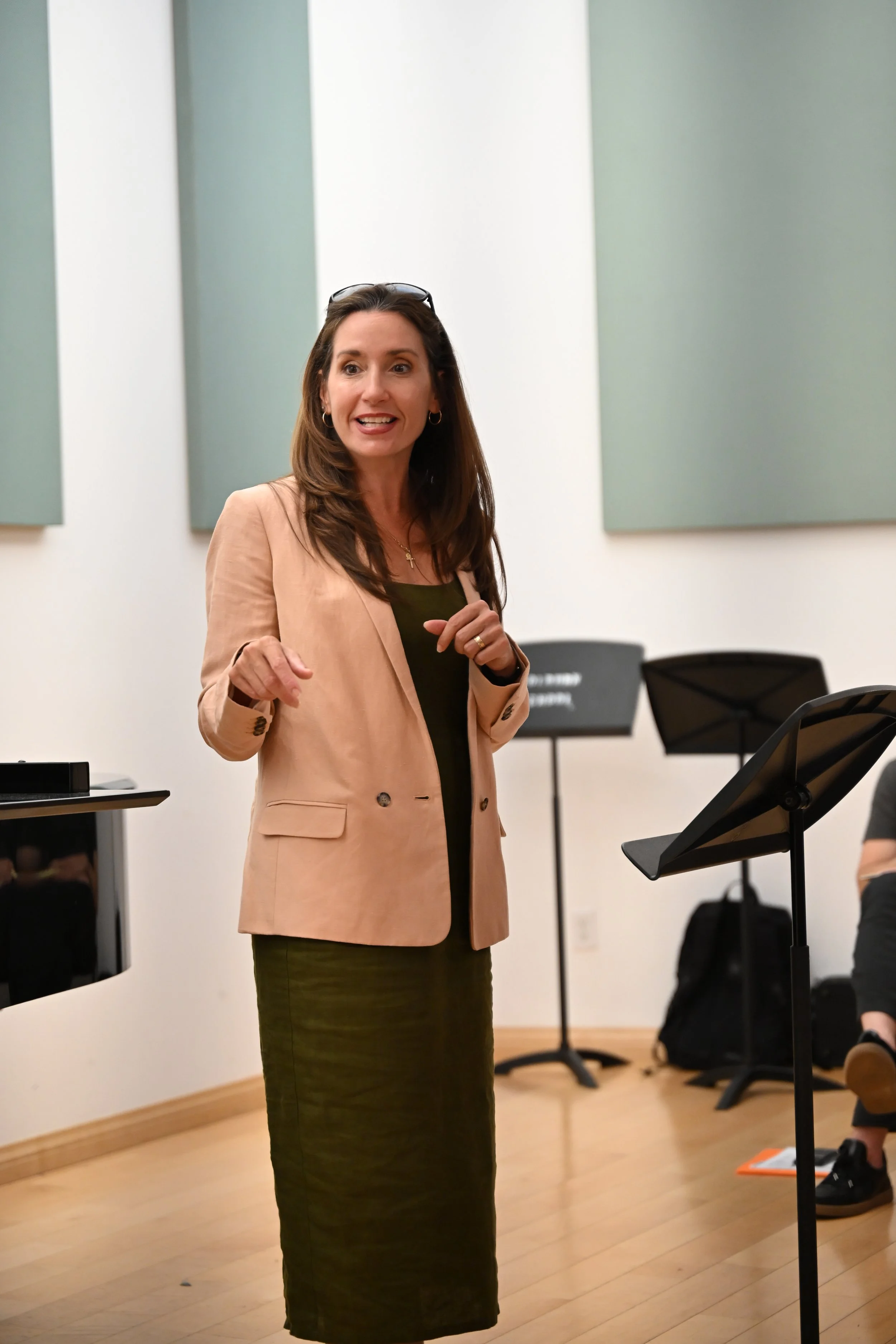 A woman with long brown hair, wearing a tan blazer and a dark green dress, standing in front of a music stand in a room with soundproofing panels on the walls, speaking or presenting to an audience.