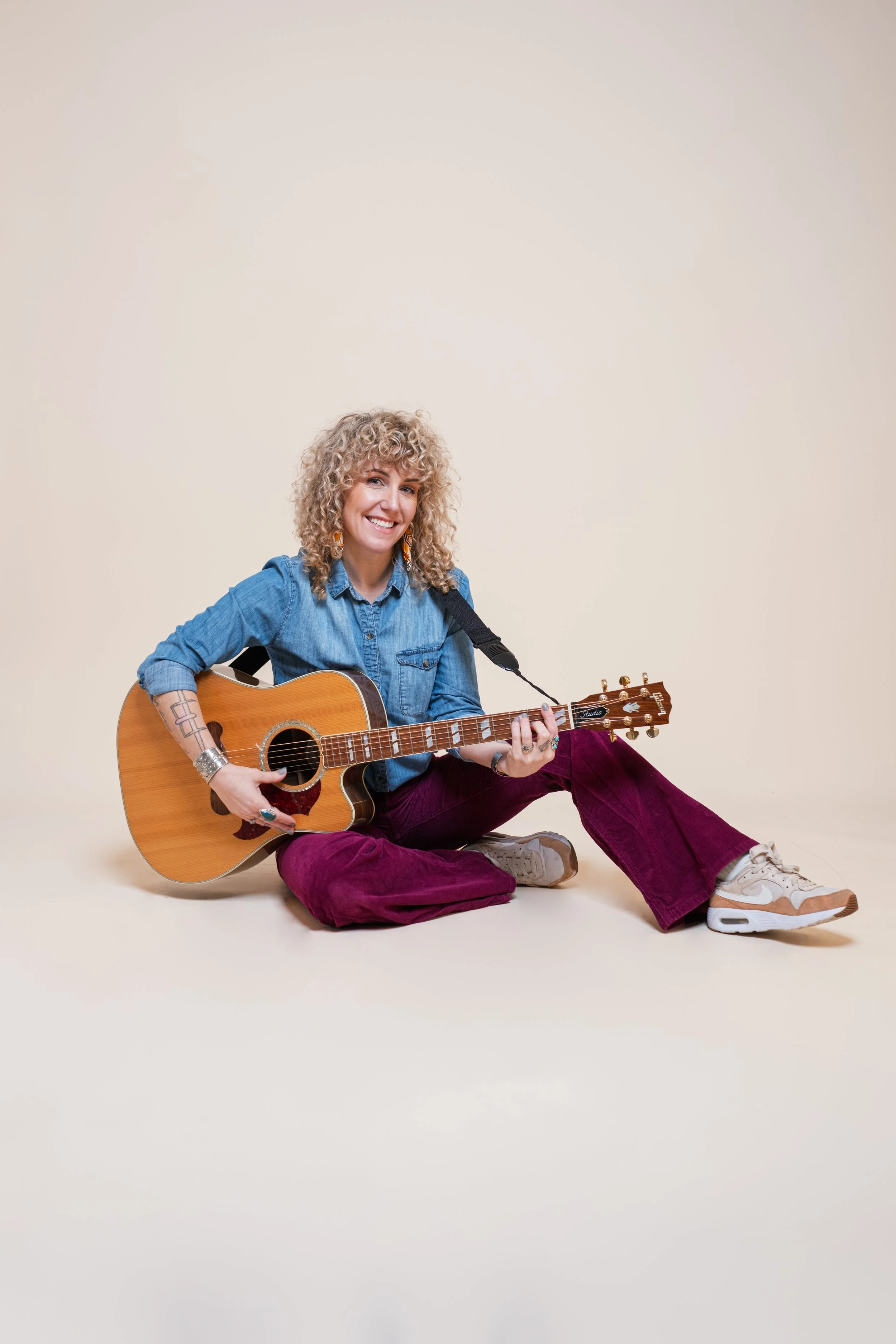Curly haired woman sits casually on the floor wearing a blue chambray button up shirt and magenta corduroy bellbottoms while strumming an acoustic guitar.