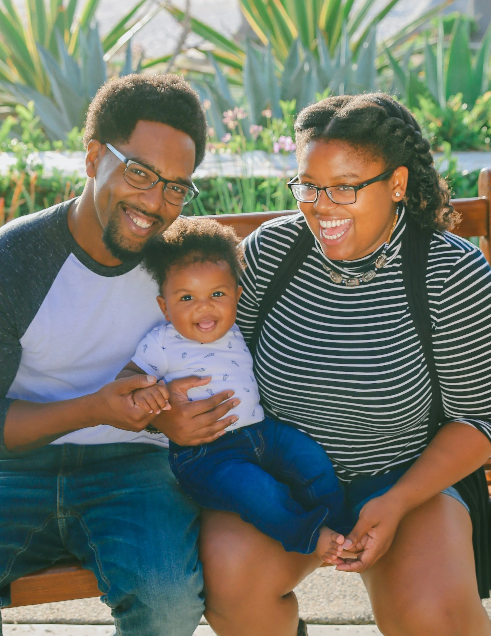 A happy family of three, including a smiling man, woman, and toddler, sitting on a bench outdoors with lush greenery in the background.