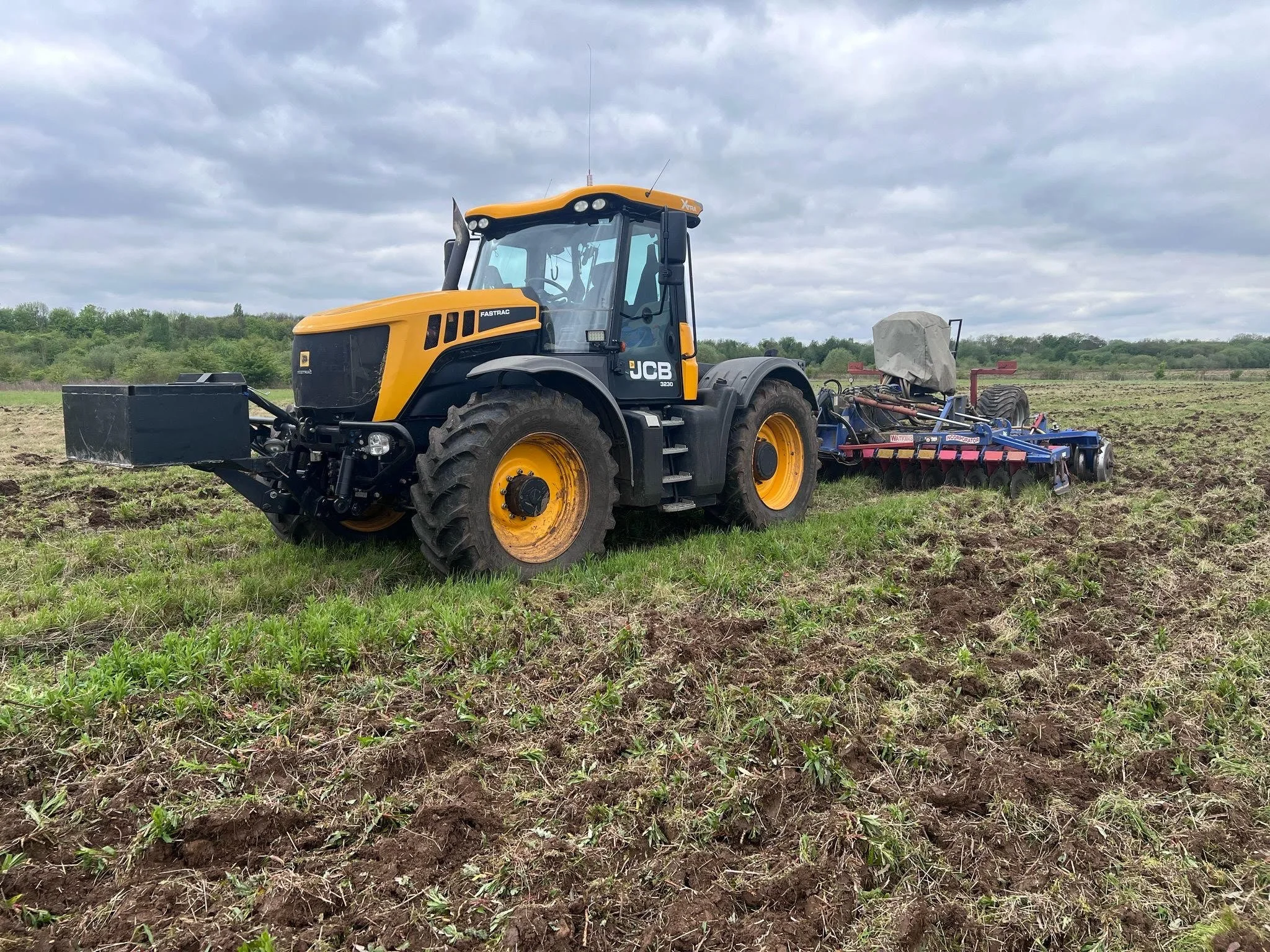 Yellow and black JCB tractor working in a large open field with cloudy sky overhead.