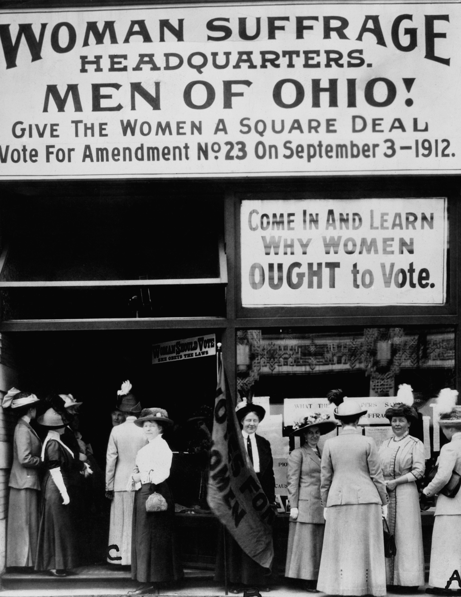 Historical black and white photograph of a women's suffrage protest outside a building. Large signs read 'Woman Suffrage Headquarters' and 'Men of Ohio! Give the women a square deal. Vote for Amendment No. 23 on September 3, 1912.' Women are dressed in early 20th-century fashion, wearing long skirts and large hats, and are gathered in front of the entrance.