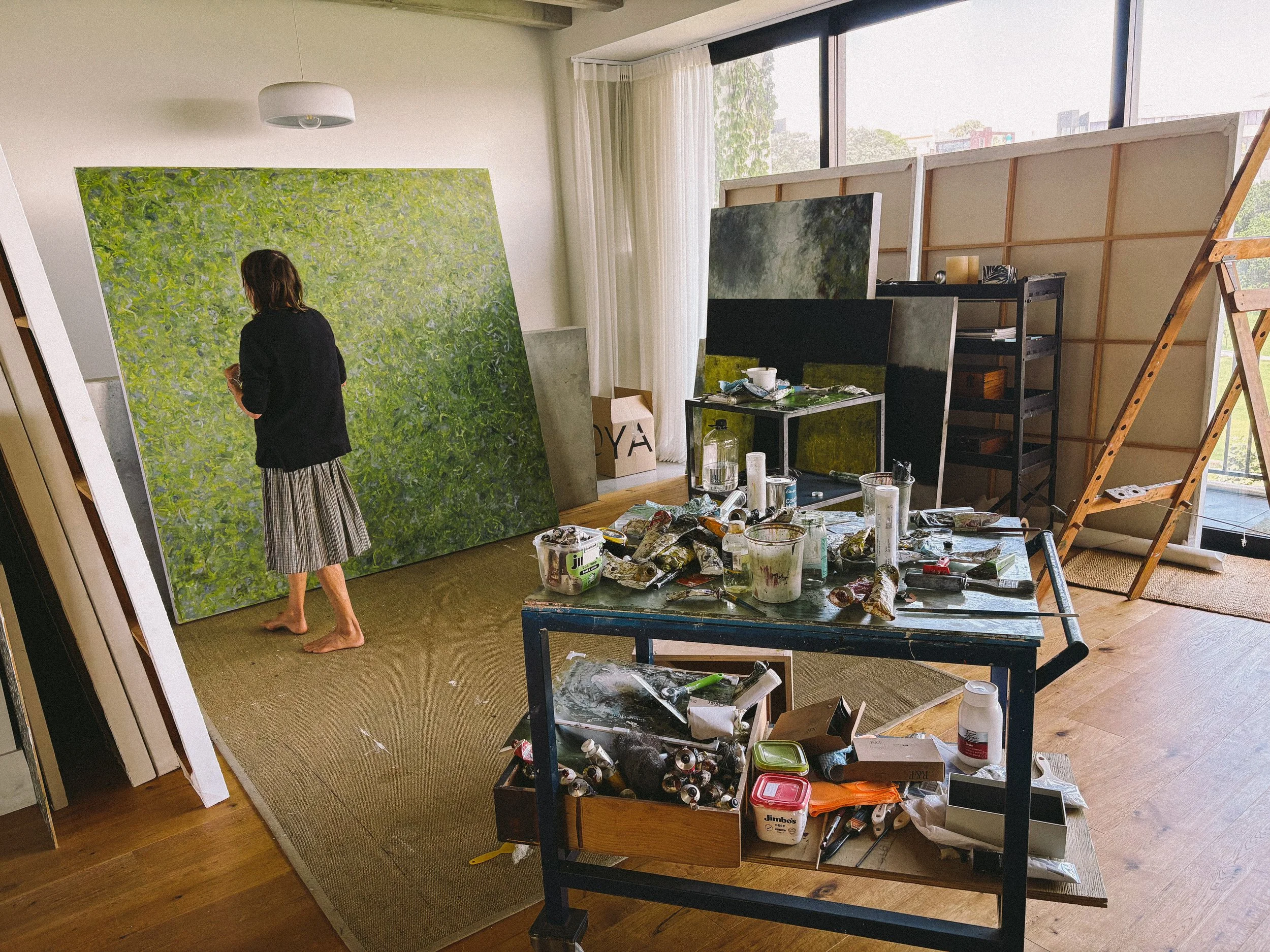 An artist's studio with a person painting a large green abstract artwork on the wall, surrounded by art supplies, canvases, and painting tools.