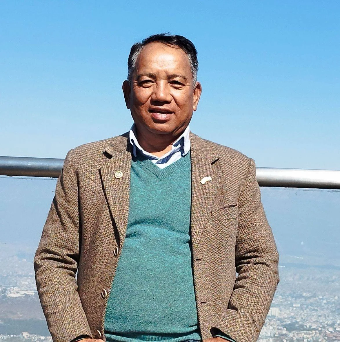 A man standing outdoors on a clear day, wearing a brown blazer, green sweater, and collared shirt, with a cityscape in the background.