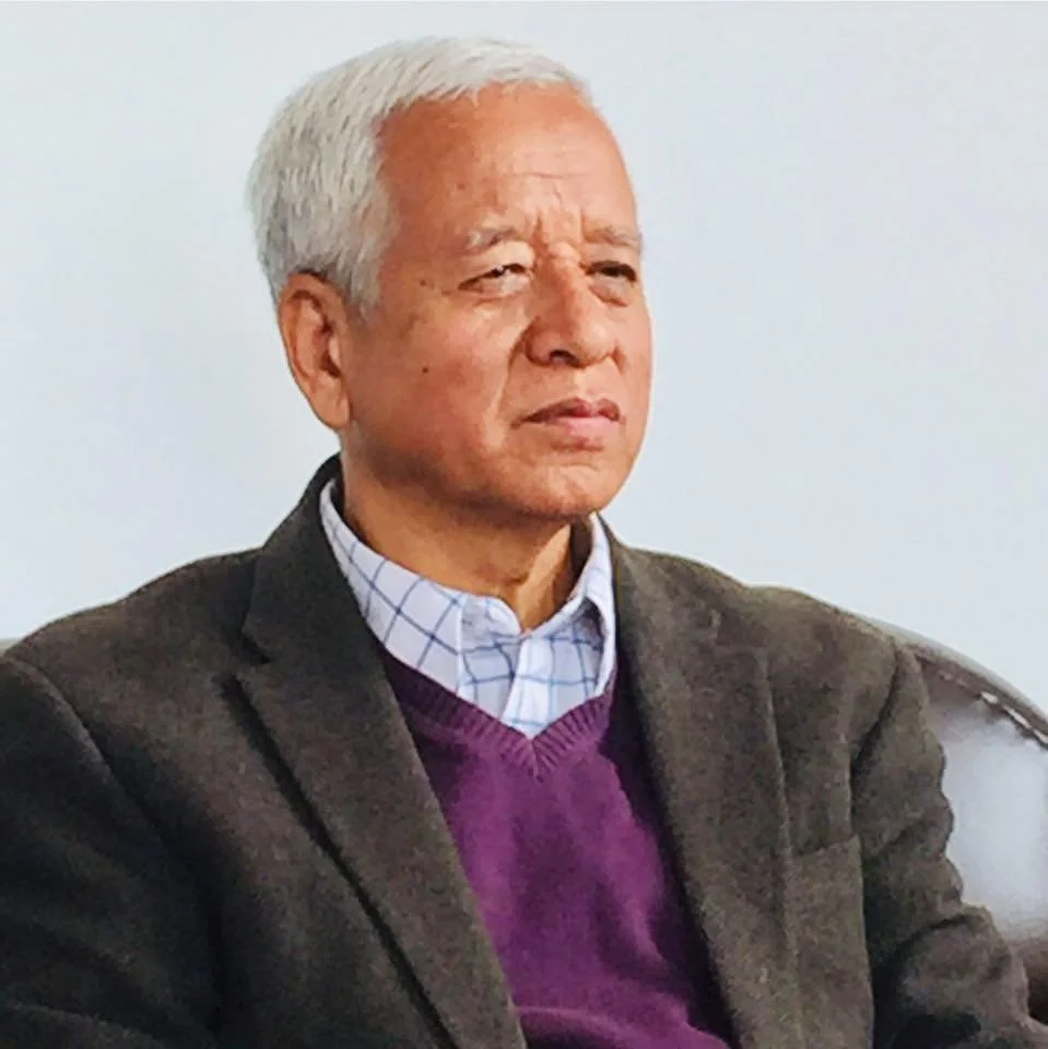 An elderly man with gray hair, wearing a brown blazer, a purple sweater, and a checkered shirt, looks serious or thoughtful.
