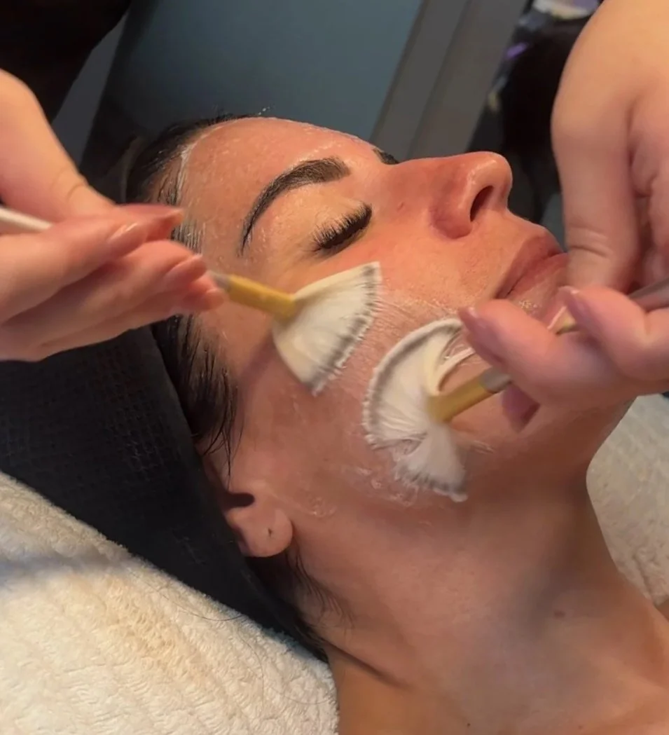 A woman is receiving a facial treatment with brushes applied to her face by two estheticians in a spa setting.