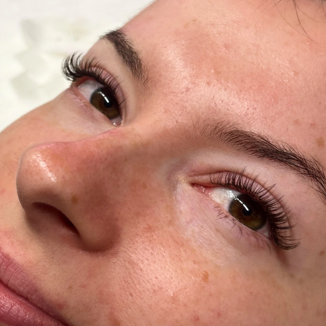Close-up of a person's face showing brown eyes, long eyelashes, eyebrows, and skin with freckles and a slightly shiny nose.