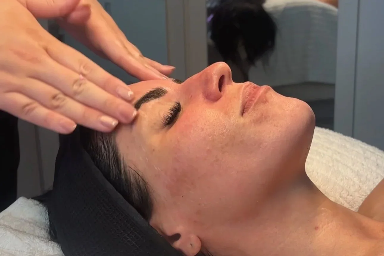 A woman receiving a facial treatment, lying down with her eyes closed while a person gently massages her face.