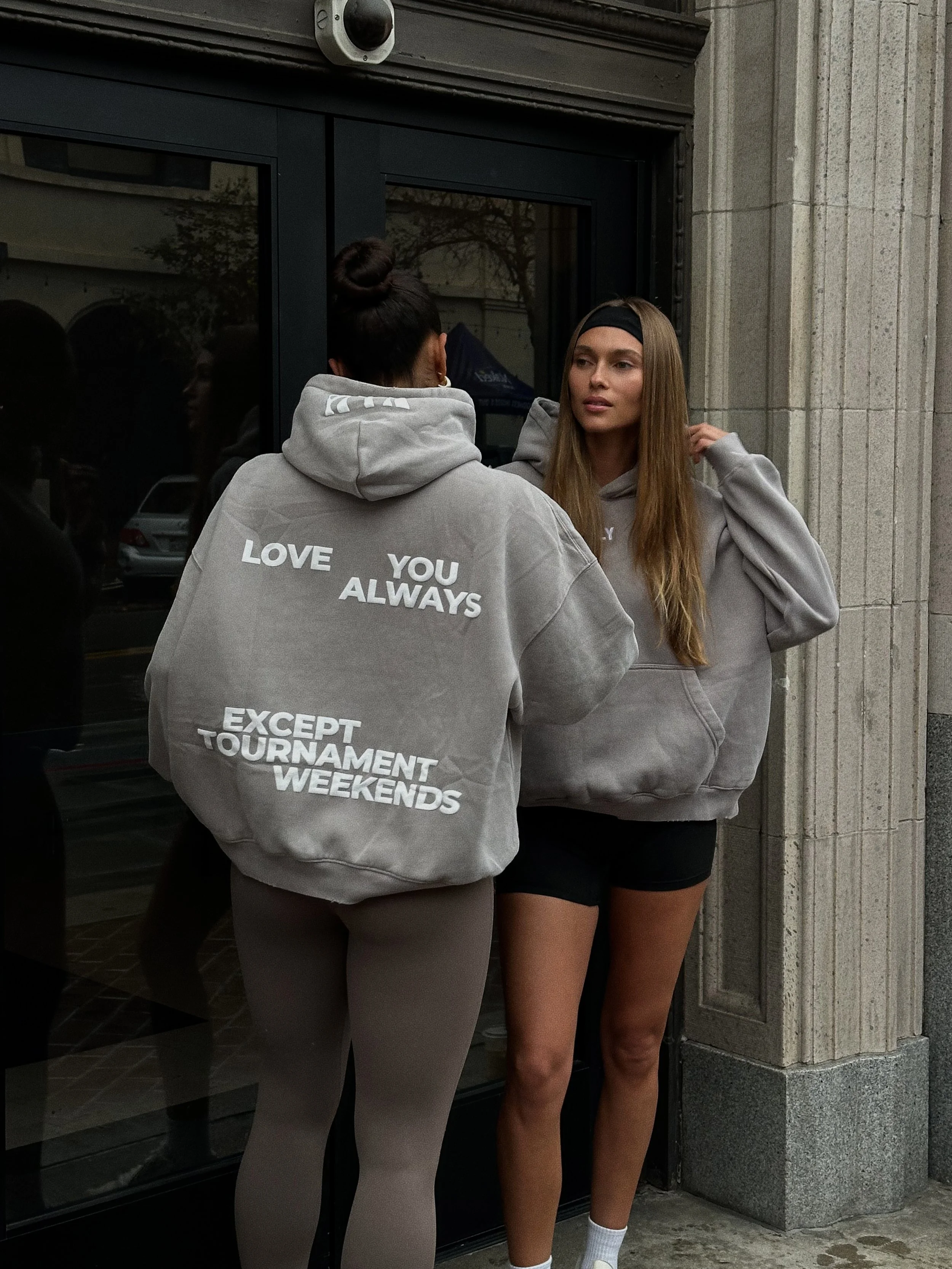 Two women conversing outside a building, both wearing gray hoodies. The woman facing away has text on her hoodie that reads 'LOVE YOU ALWAYS EXCEPT TOURNAMENT WEEKENDS.' The woman facing the camera has long hair, a headband, and is adjusting her hoodie.
