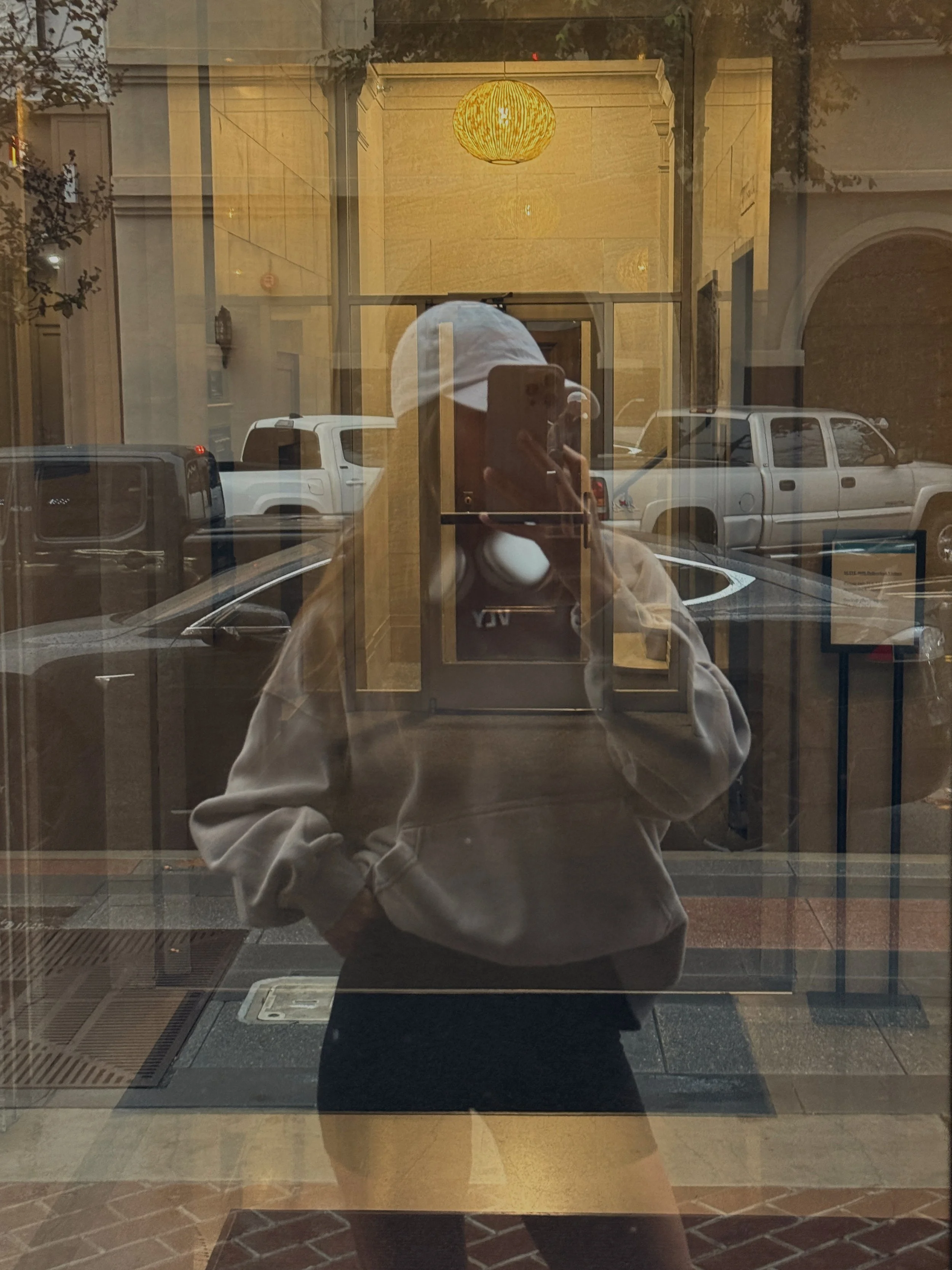 Reflection of a person taking a photo with a smartphone in a glass window. The person is wearing a grey hoodie and a white cap. The background shows parked cars and a building entrance with a yellow hanging light