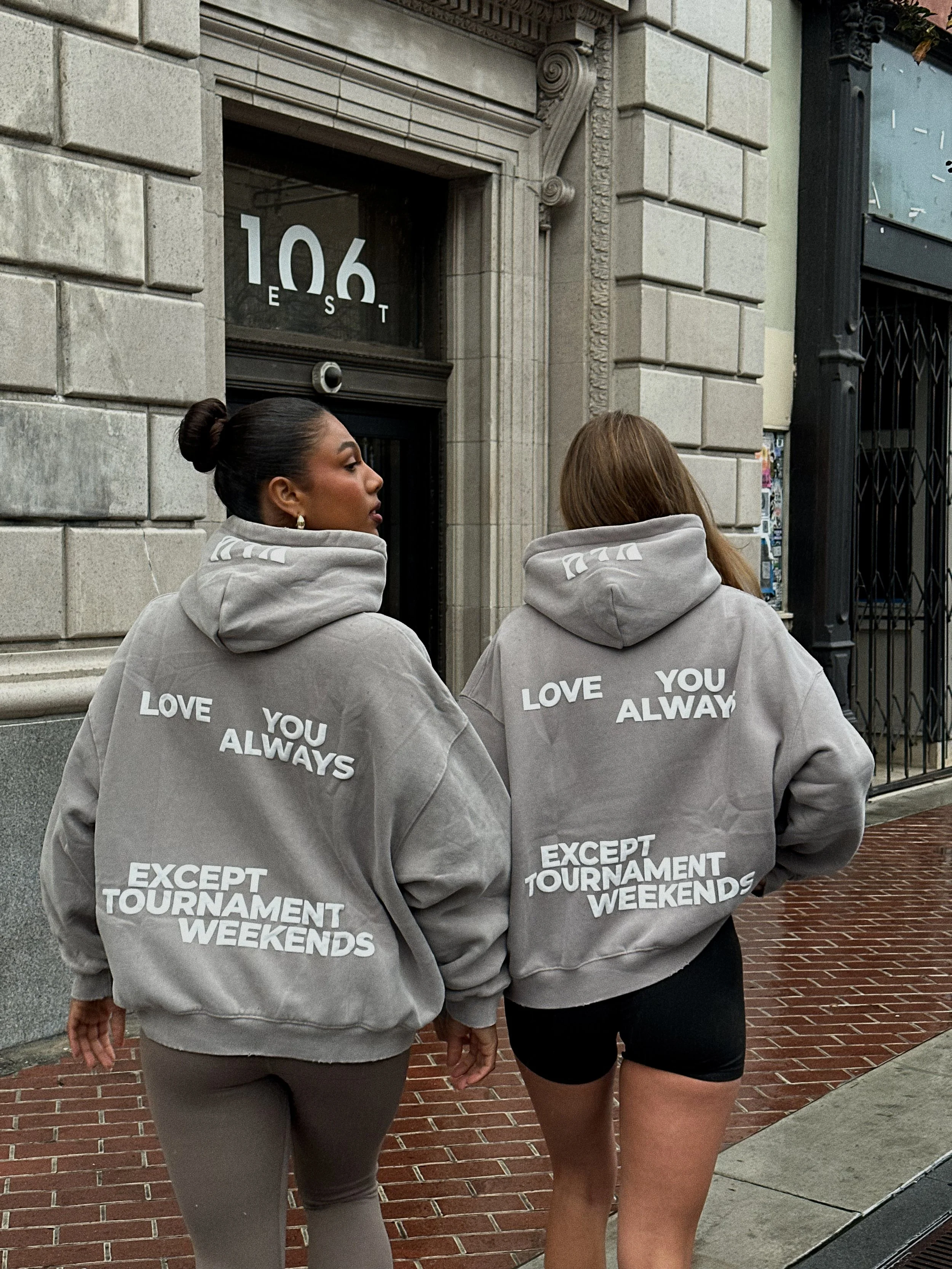 Two women walking on a city sidewalk wearing matching gray hoodies with white text. The woman on the left has dark hair in a bun and is wearing beige pants, while the woman on the right has light brown hair and is wearing black shorts. The hoodie on the left reads, "LOVE YOU ALWAYS," and the hoodie on the right reads, "LOVE YOU ALWAYS EXCEPT TOURNAMENT WEEKENDS." They are walking past a building with the address "106 E ST" on the door.