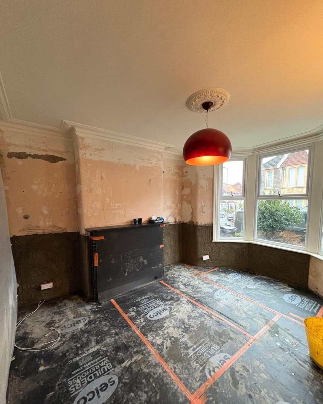 Room under renovation with stripped walls, partially removed wallpaper, and a black panel against the wall. The floor is covered with protective sheets, and there is a large window letting in natural light.