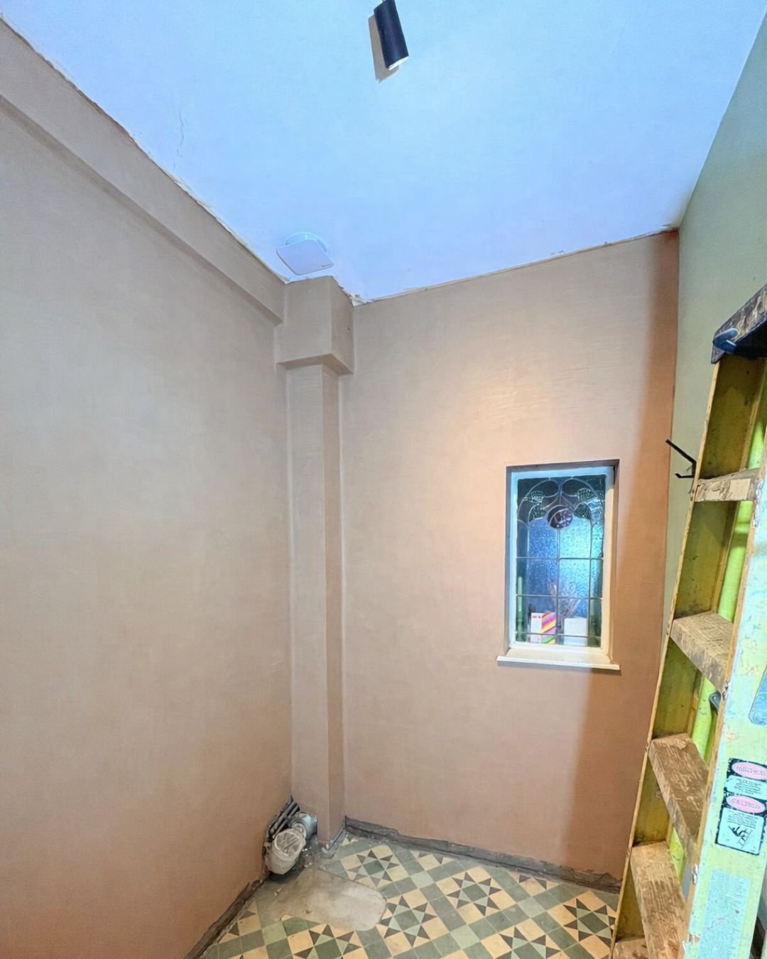 A small room under renovation with beige and green walls, a patterned tile floor, a small window with decorative glass, and a ladder on the right side.