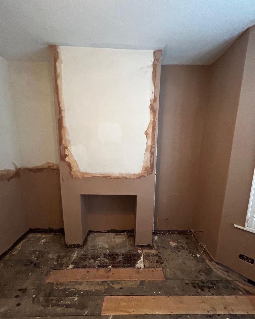 Room with wall partially torn down, exposing drywall and framing, with damaged flooring and a small rectangular opening in the wall.
