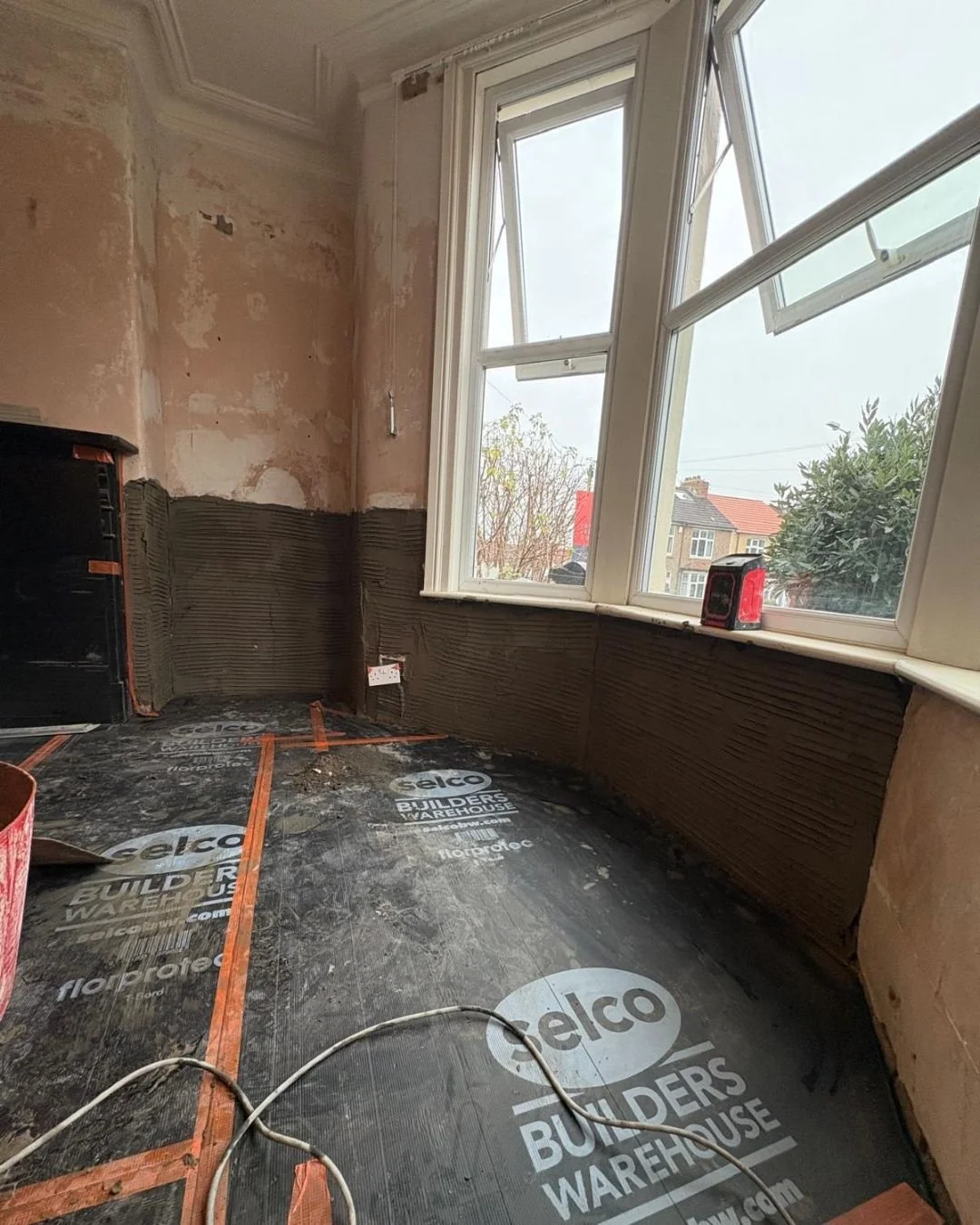 Room under renovation with partially painted walls, a large window, and a black waterproof floor covering marked "Celco Builders Warehouse." An electrical cable and a red spirit level are on the floor, and there is a black source of heat or power on 