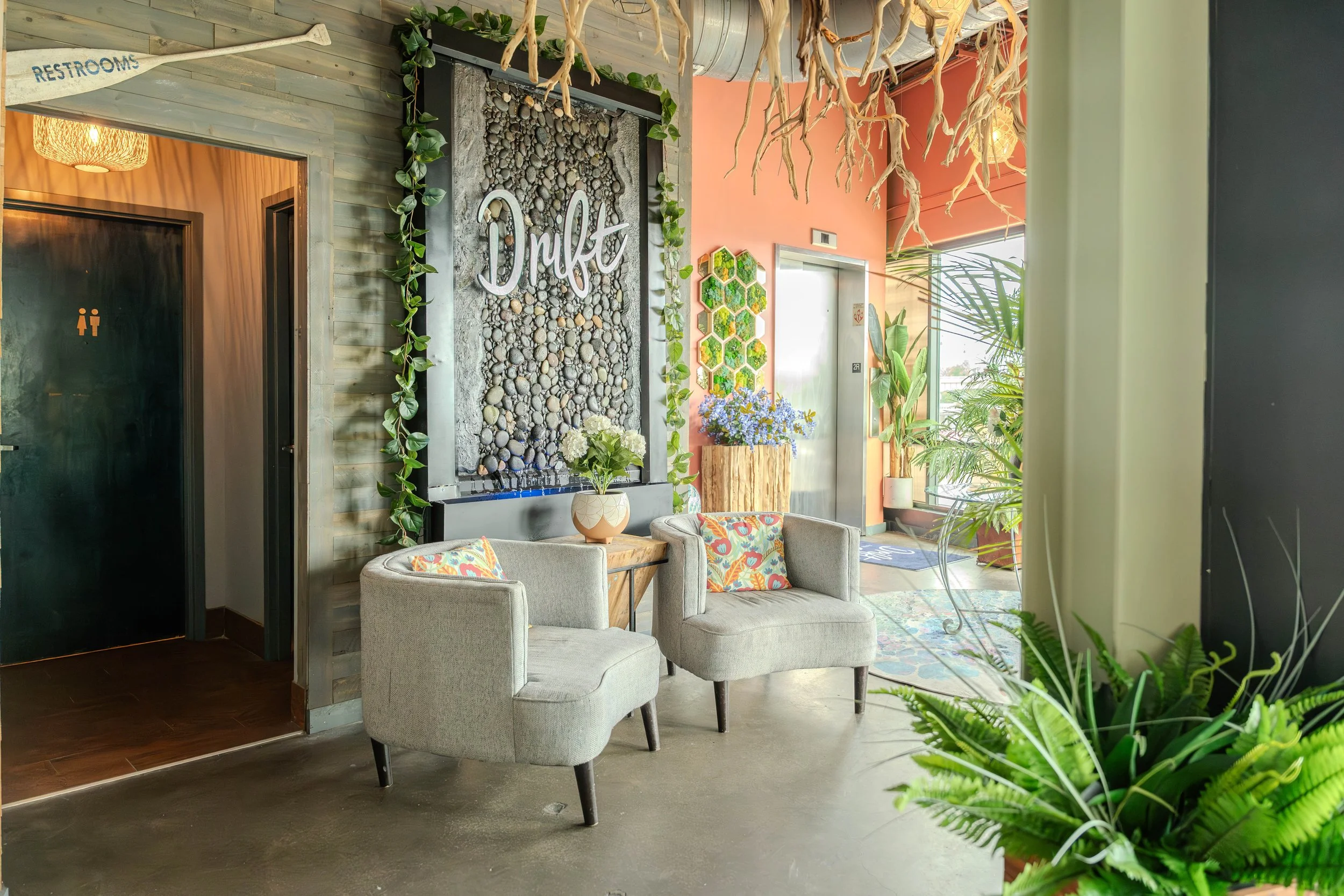A cozy lobby area with two beige armchairs, colorful throw pillows, and a small wooden side table with a vase of white flowers. Behind the chairs is a wall decorated with a large pebble art piece with the word 'Drift' in white cursive. The space features indoor plants, a hanging driftwood chandelier, a pink wall with hexagonal green planters, and an elevator door. To the left, an entrance to restrooms with a sign overhead.