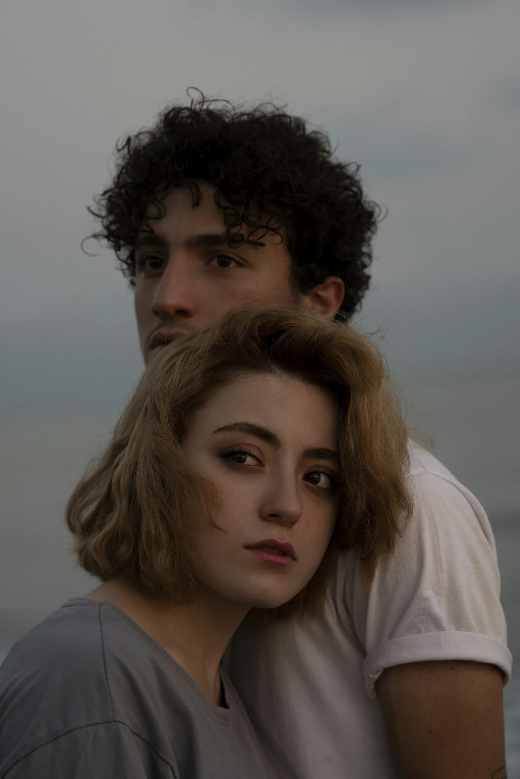 A young man with curly dark hair and a young woman with short, wavy, reddish hair standing close together outdoors, with overcast sky in the background.