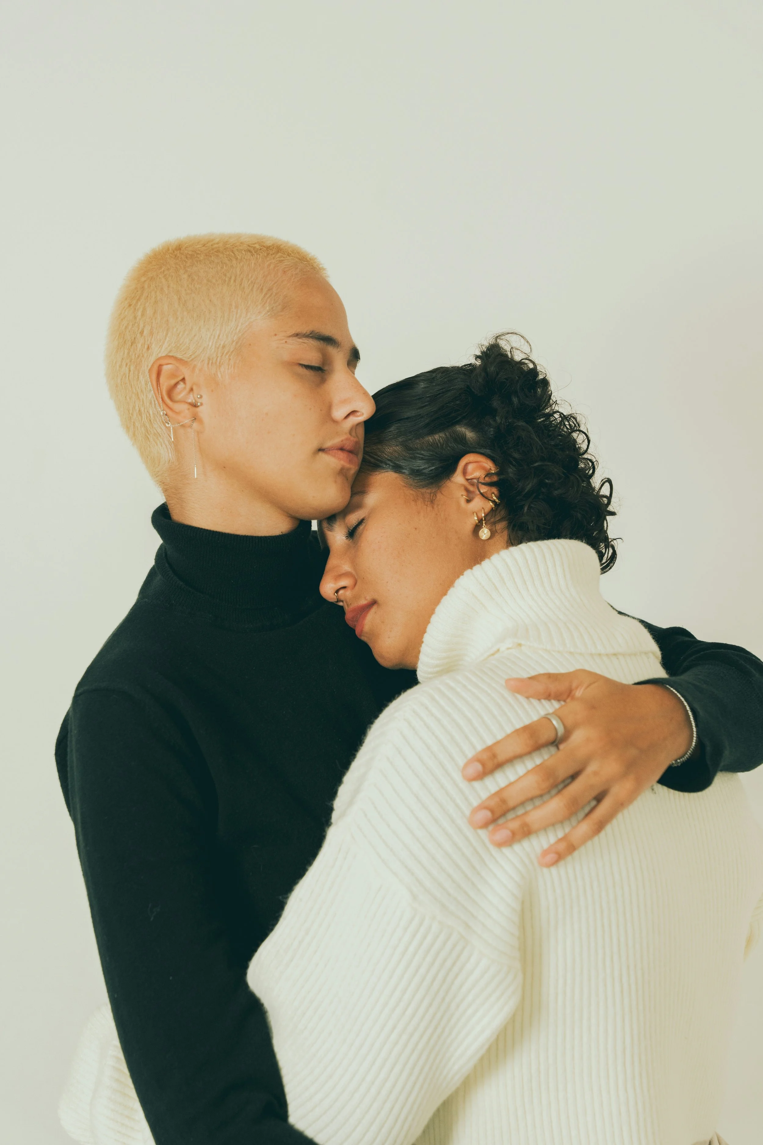 A close-up of two women embracing, with their eyes closed and their foreheads touching. One woman has short, platinum blonde hair and wears a black turtleneck; the other has dark, curly hair and wears a white turtleneck.