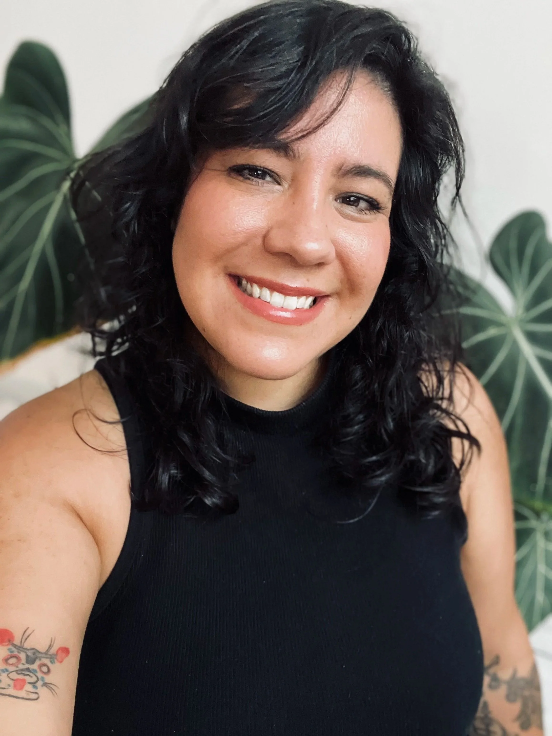 A woman with black, curly hair smiling at the camera, wearing a black sleeveless top with a tattoo visible on her arm, and standing in front of large green leaves.