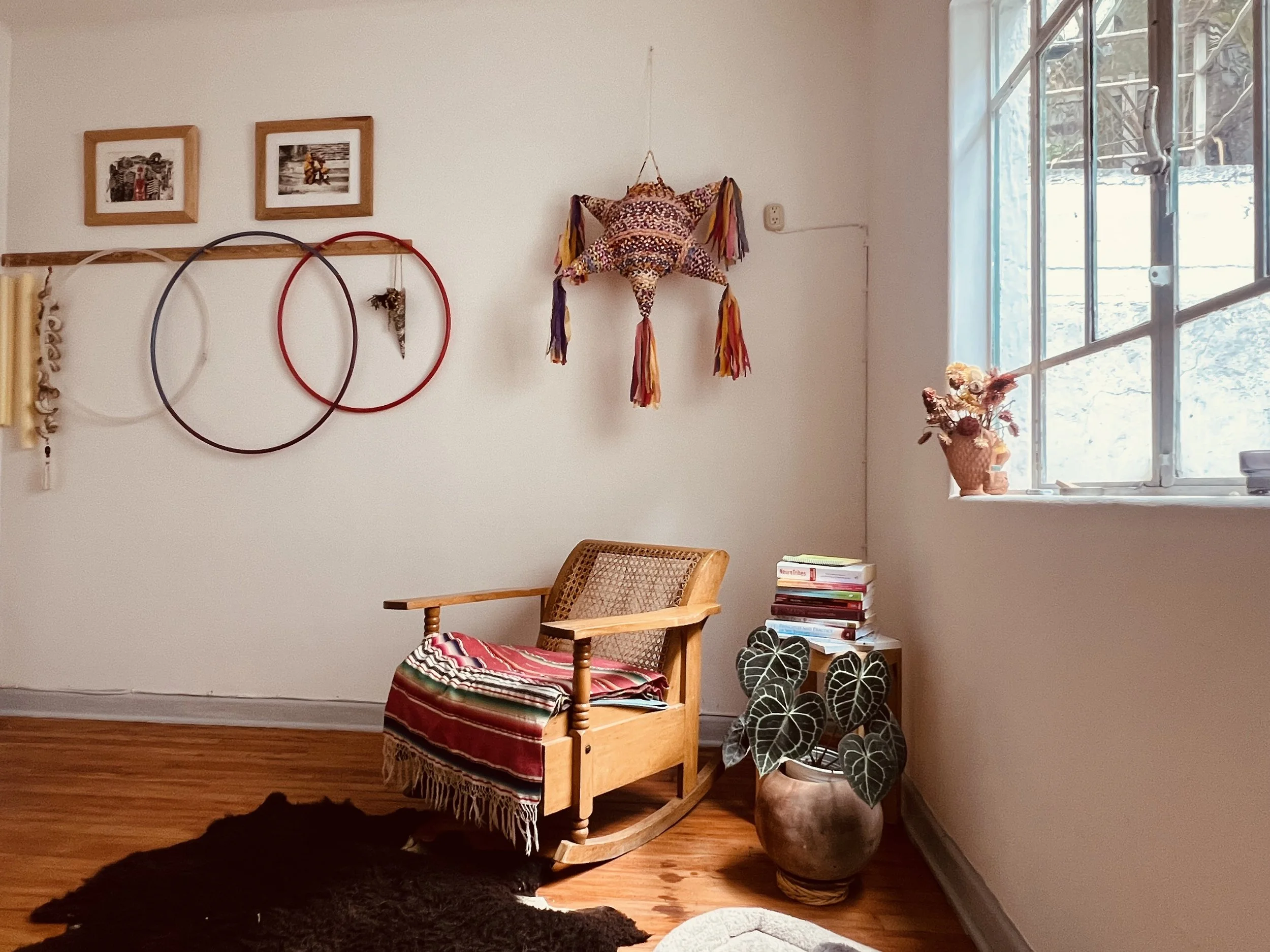 Interior of a cozy room with a wooden rocking chair covered with a patterned blanket, a stack of books beside it, a potted plant on the floor, and wall decorations including framed photos, hula hoops, and a star-shaped tapestry, with sunlight coming through a large window.