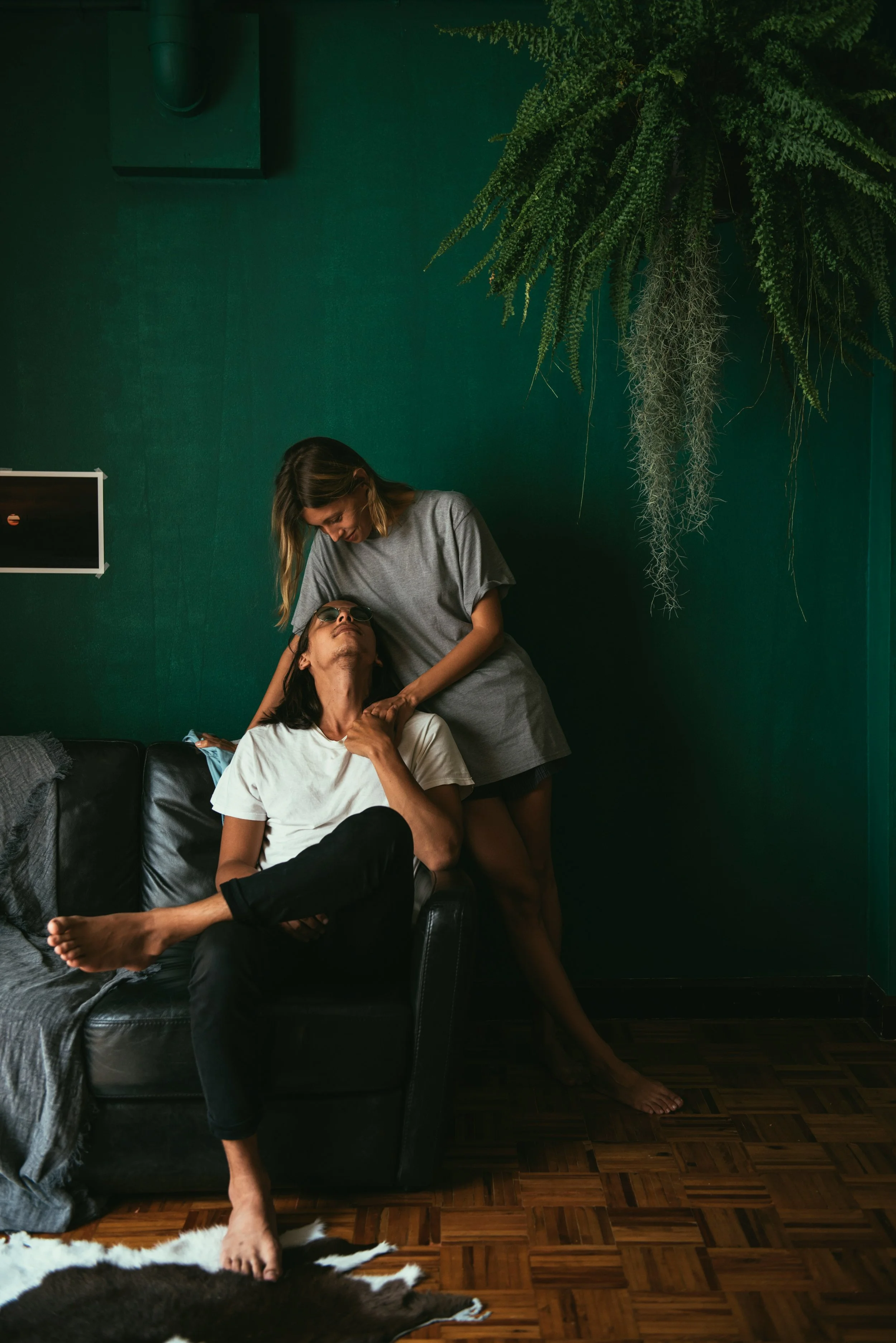 Two women interact in a dimly lit room with green walls and wooden flooring. One woman is sitting on a black leather couch with her head tilted back, wearing sunglasses and a white T-shirt. The other woman stands beside her, leaning over and touching her neck gently. There are hanging green plants on the wall, and a small flat-screen TV or monitor is partially visible on the wall.