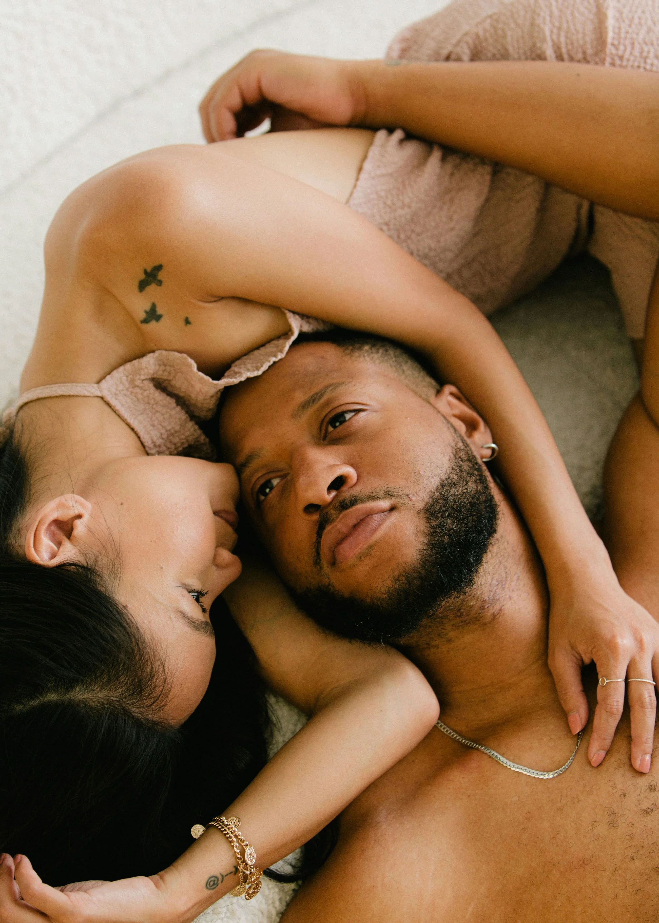 A man and woman lying on the floor close together, with the woman resting her head on the man's shoulder and face close to his, looking into each other's eyes. The woman is touching the man's chest and the man has his arm around her.