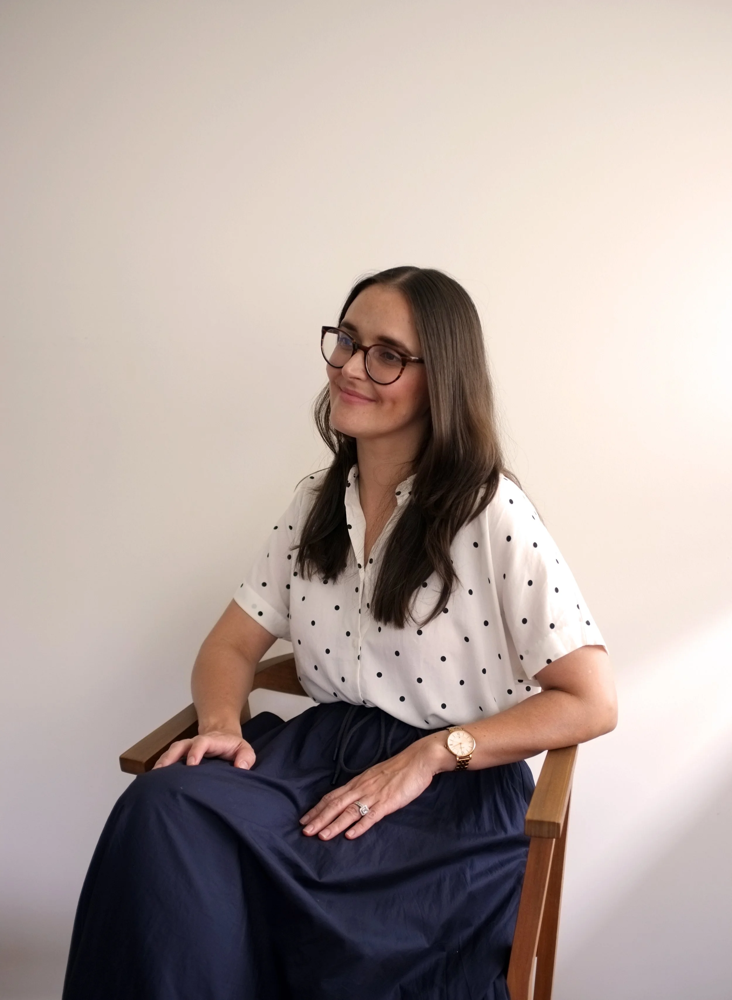 A woman with long dark hair, wearing glasses, a white polka dot shirt, and a navy skirt, sitting on a wooden chair against a plain white background. She is smiling softly and looking slightly to the side.