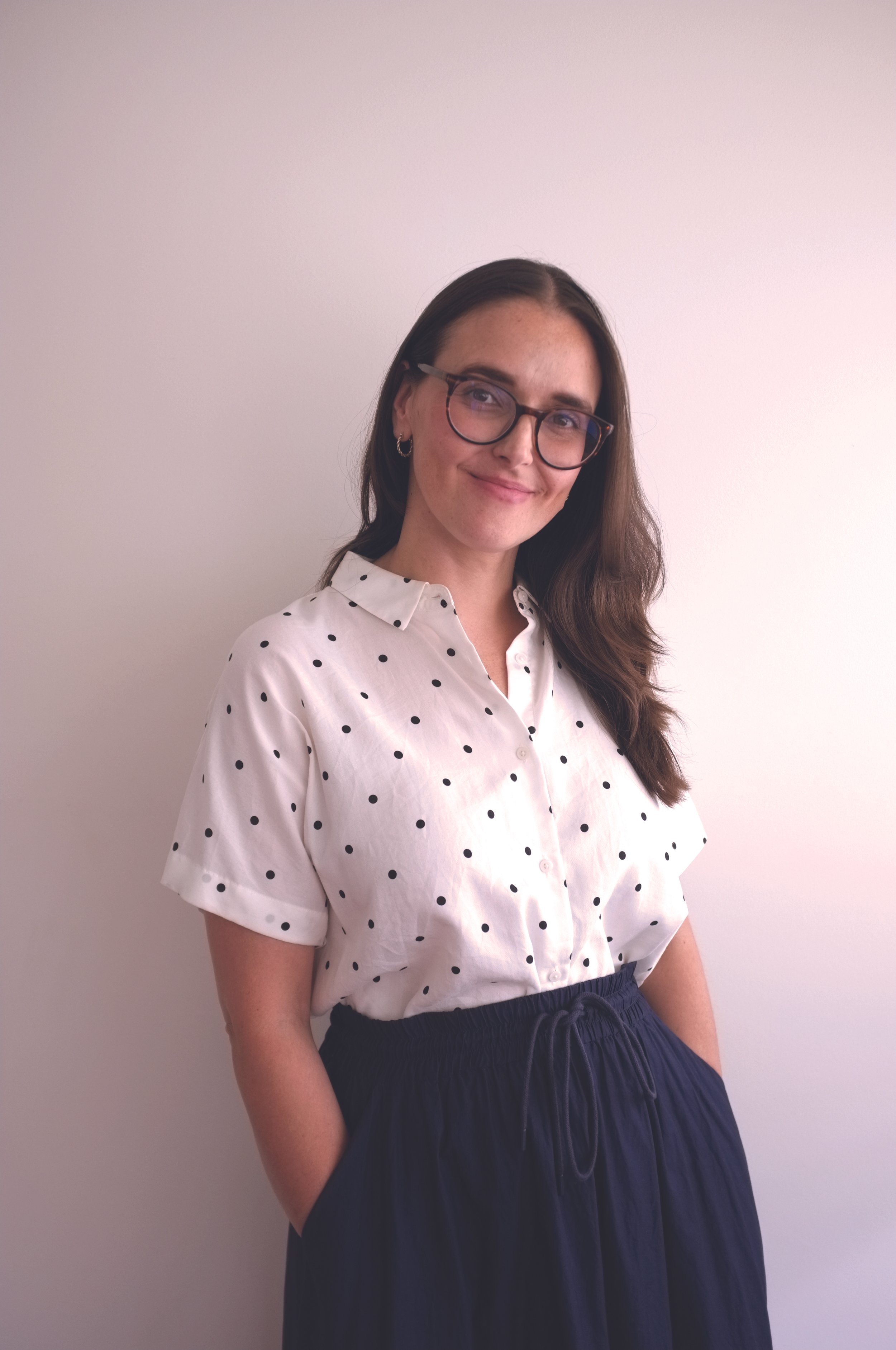 A woman with glasses and earrings, wearing a white polka dot shirt and navy blue skirt, smiling and standing against a plain light-colored wall.