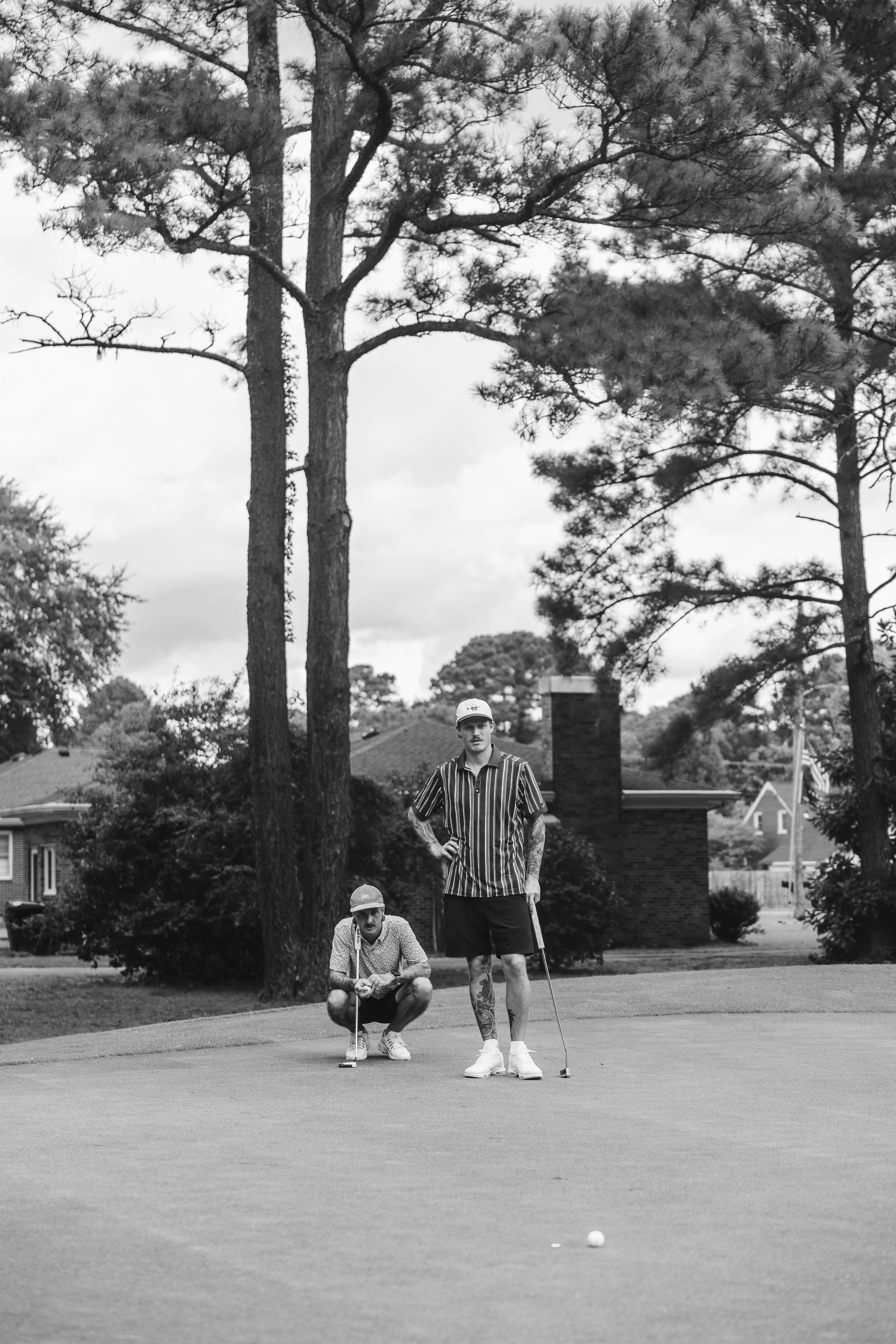 Two men playing golf on a golf course, with one standing and one crouching near the putting green, trees and houses in the background, black and white photograph.