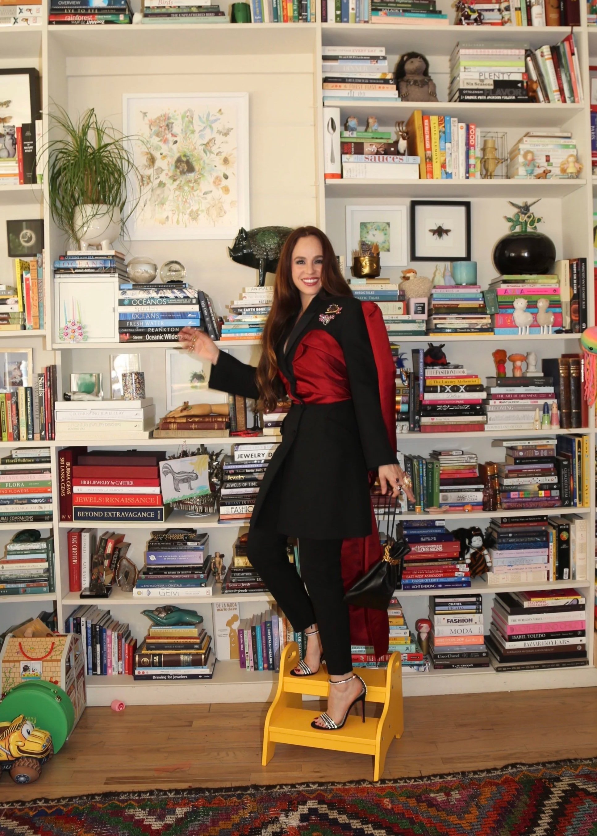 A woman with long brown hair standing in a room filled with bookshelves and colorful and fun decorative objects..She is smiling, wearing a black and red outfit with high heels. The background includes many books, art  and curiosities.