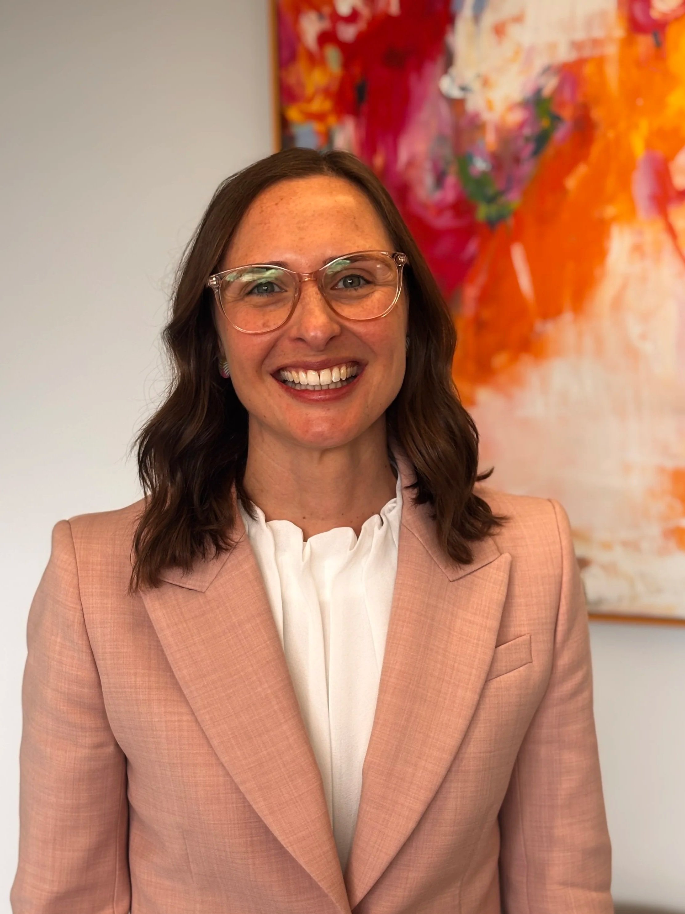 A woman with shoulder-length brown hair, wearing glasses, a white blouse, and a peach-colored blazer, smiling in front of a colorful abstract painting.