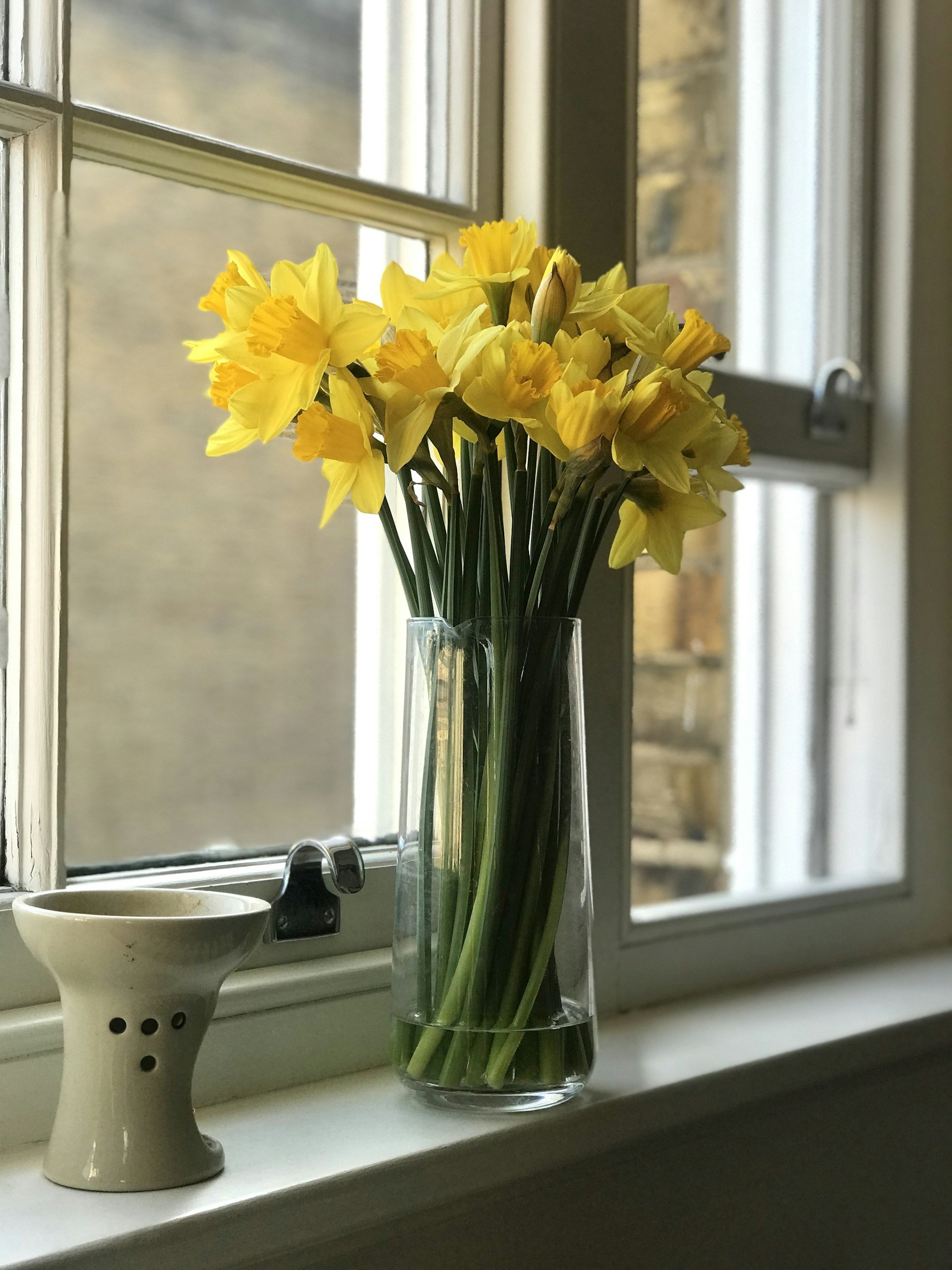 A vase of yellow daffodils on a windowsill with a white ceramic candle holder nearby.