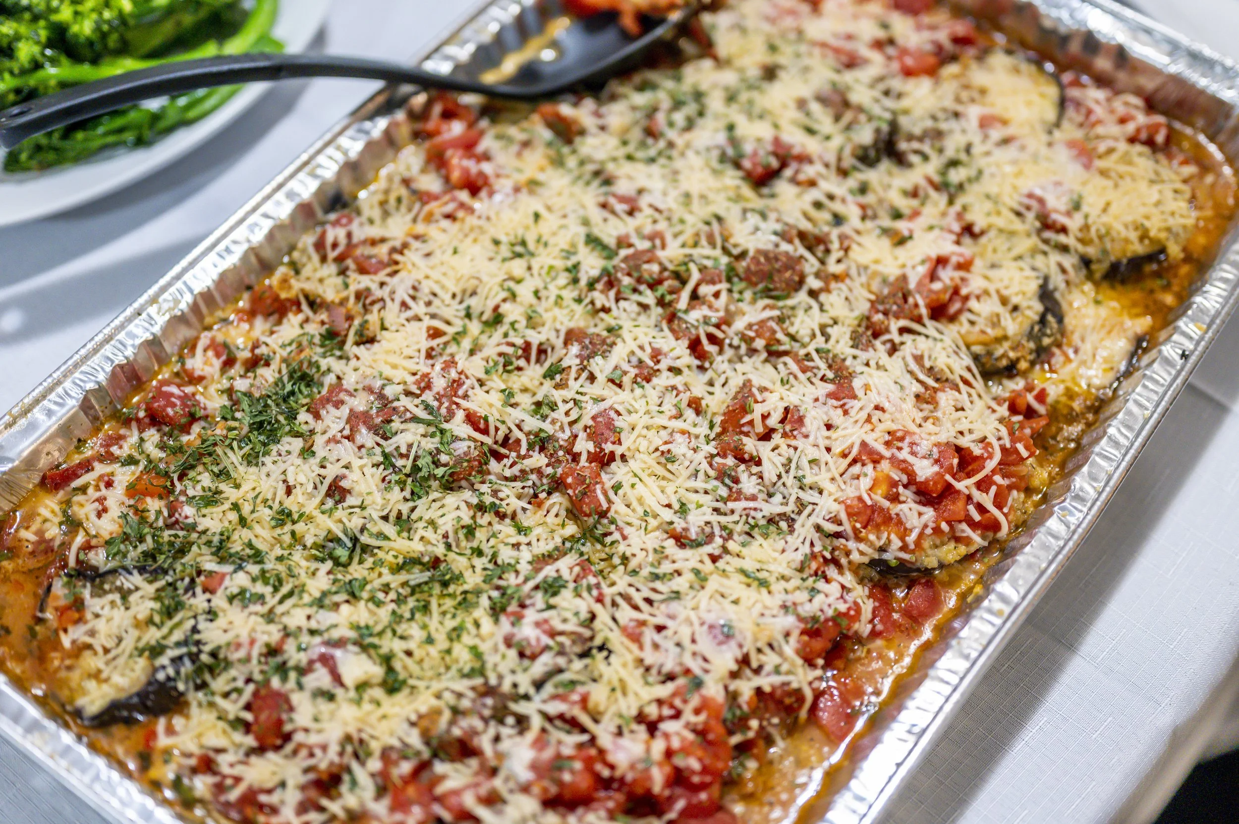 Baked eggplant parmesan topped with shredded cheese and chopped herbs in a foil tray.