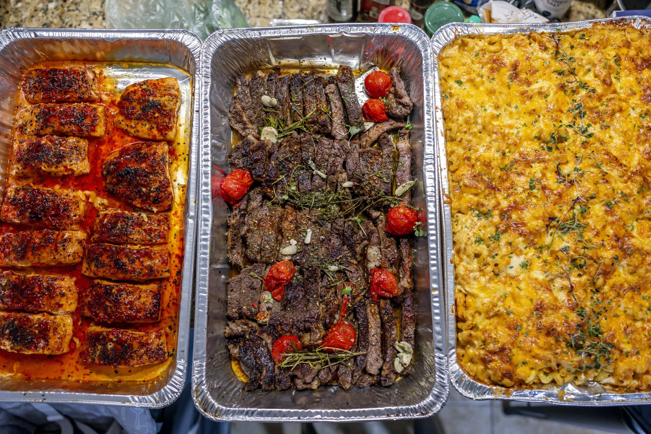 Three aluminum trays filled with grilled chicken wings, grilled steak with cherry tomatoes and garlic, and baked cheesy pasta with herbs.