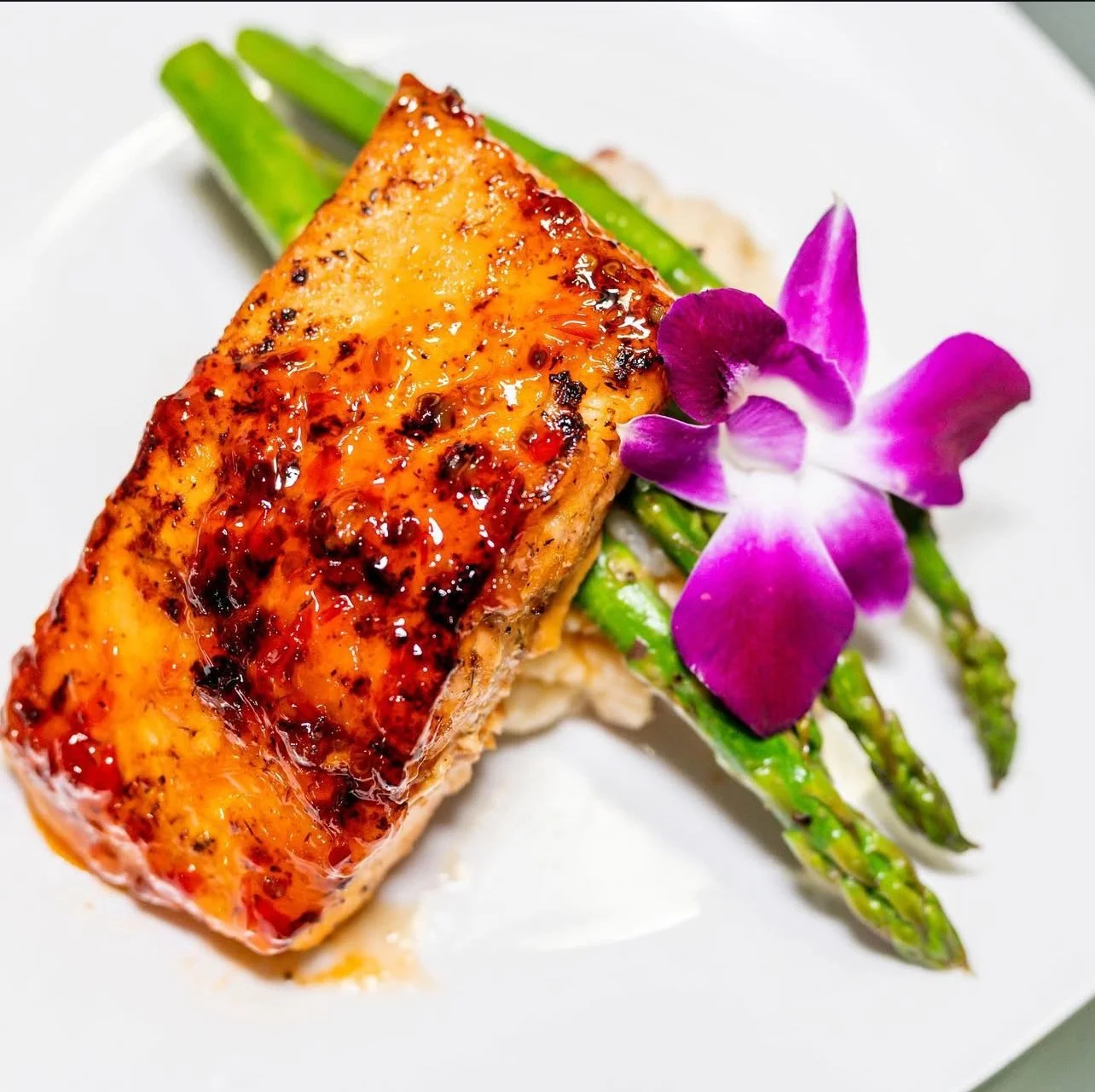 A plated meal featuring a glazed salmon fillet, grilled asparagus spears, and an edible purple orchid flower for garnish.