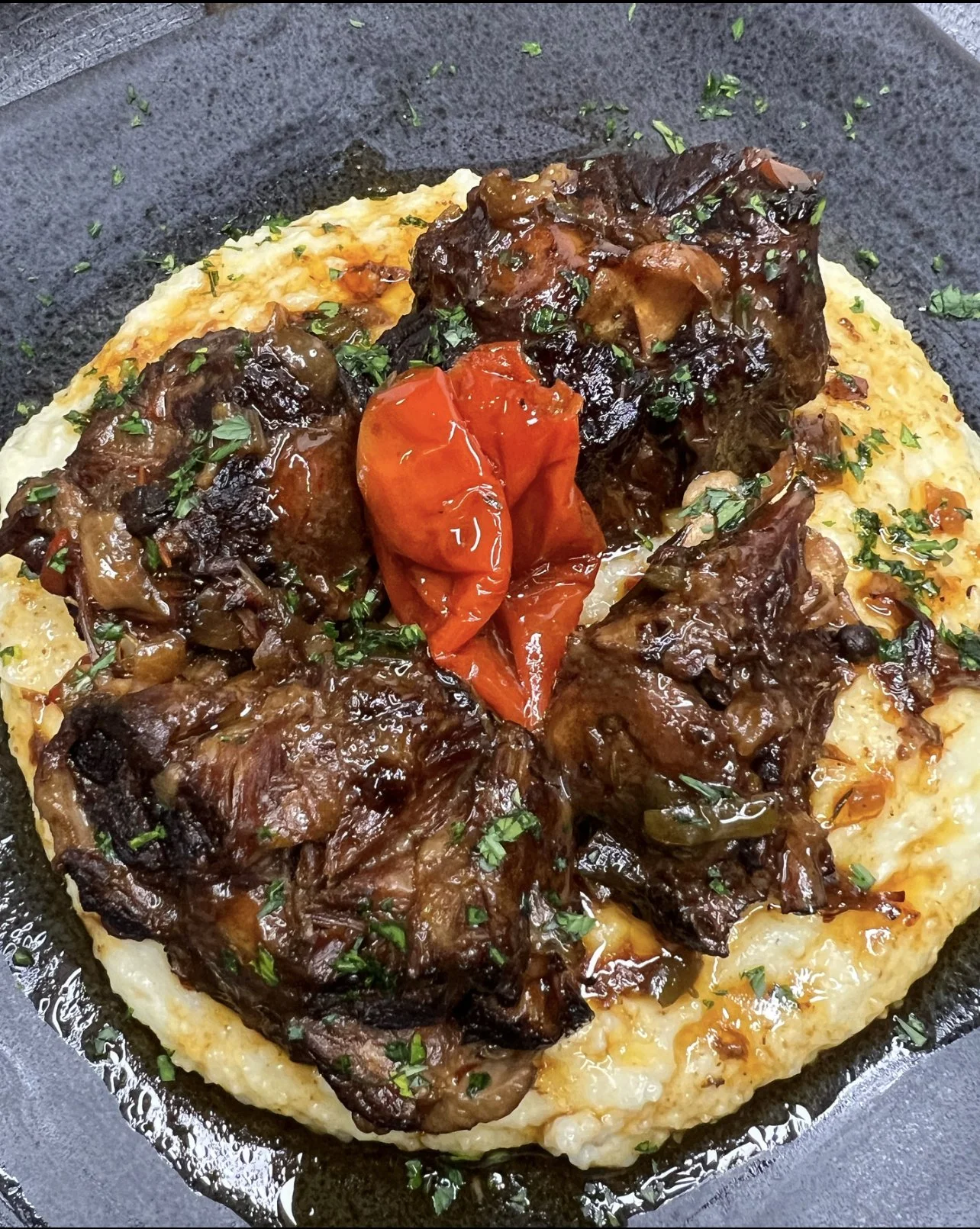 A dish with a serving of creamy mashed potatoes topped with braised beef, roasted cherry tomatoes, and chopped herbs, served on a dark plate.