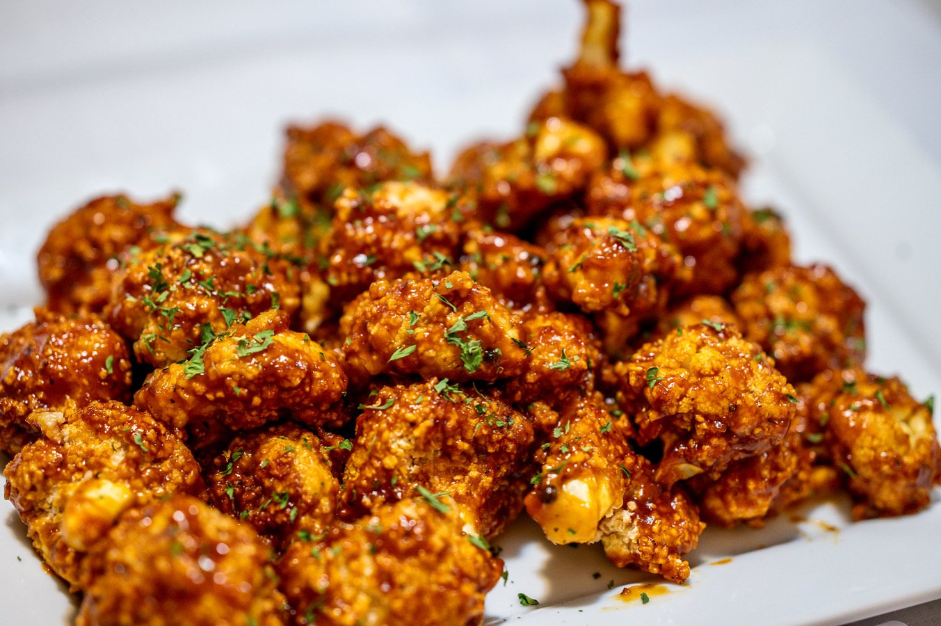 Close-up of crispy fried chicken bites glazed in sauce, garnished with chopped herbs on a white platter.