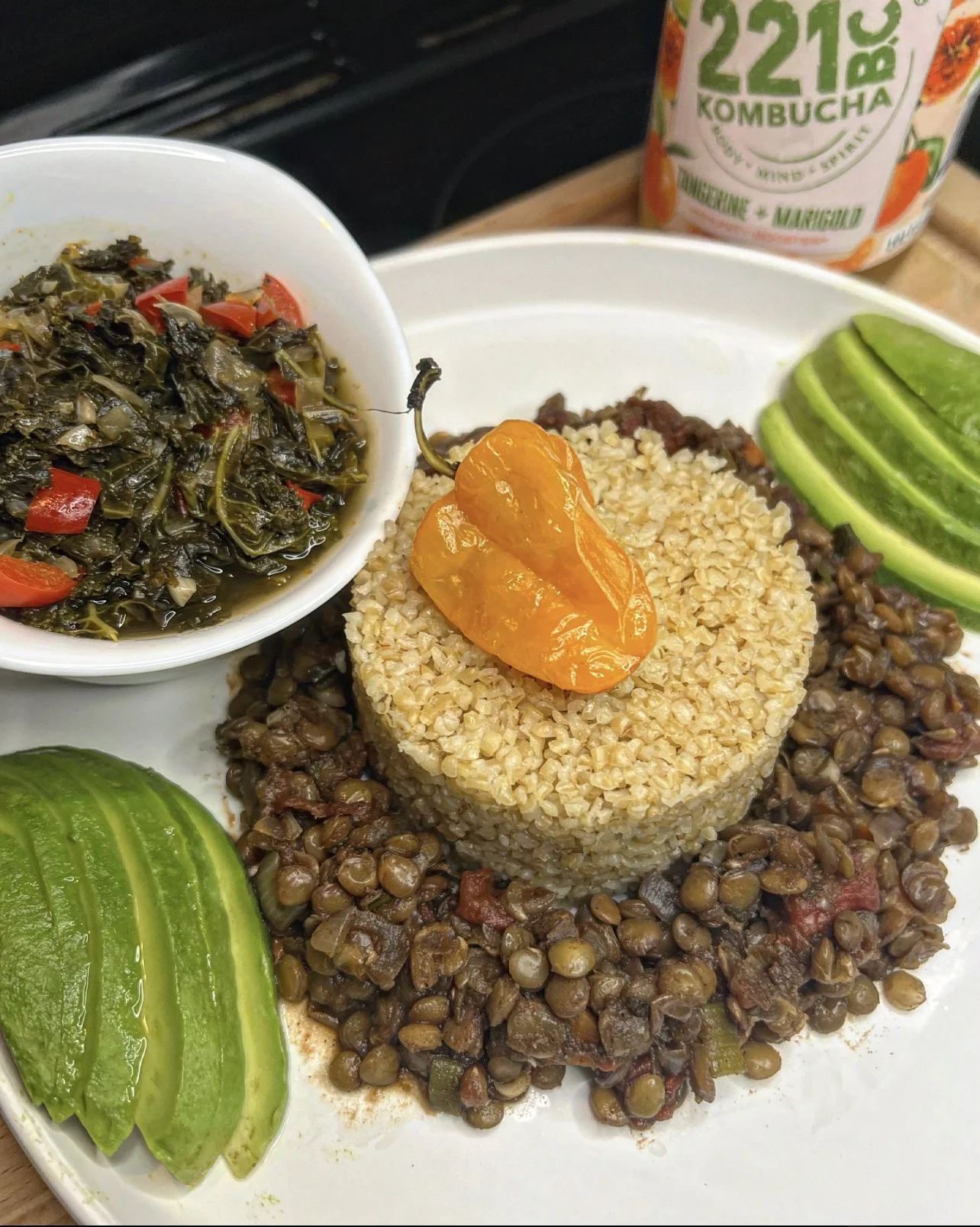 A plate of Mexican-style rice with lentils, sliced avocado, a yellow chili pepper, and cooked greens with red bell pepper, along with an orange kombucha bottle in the background.
