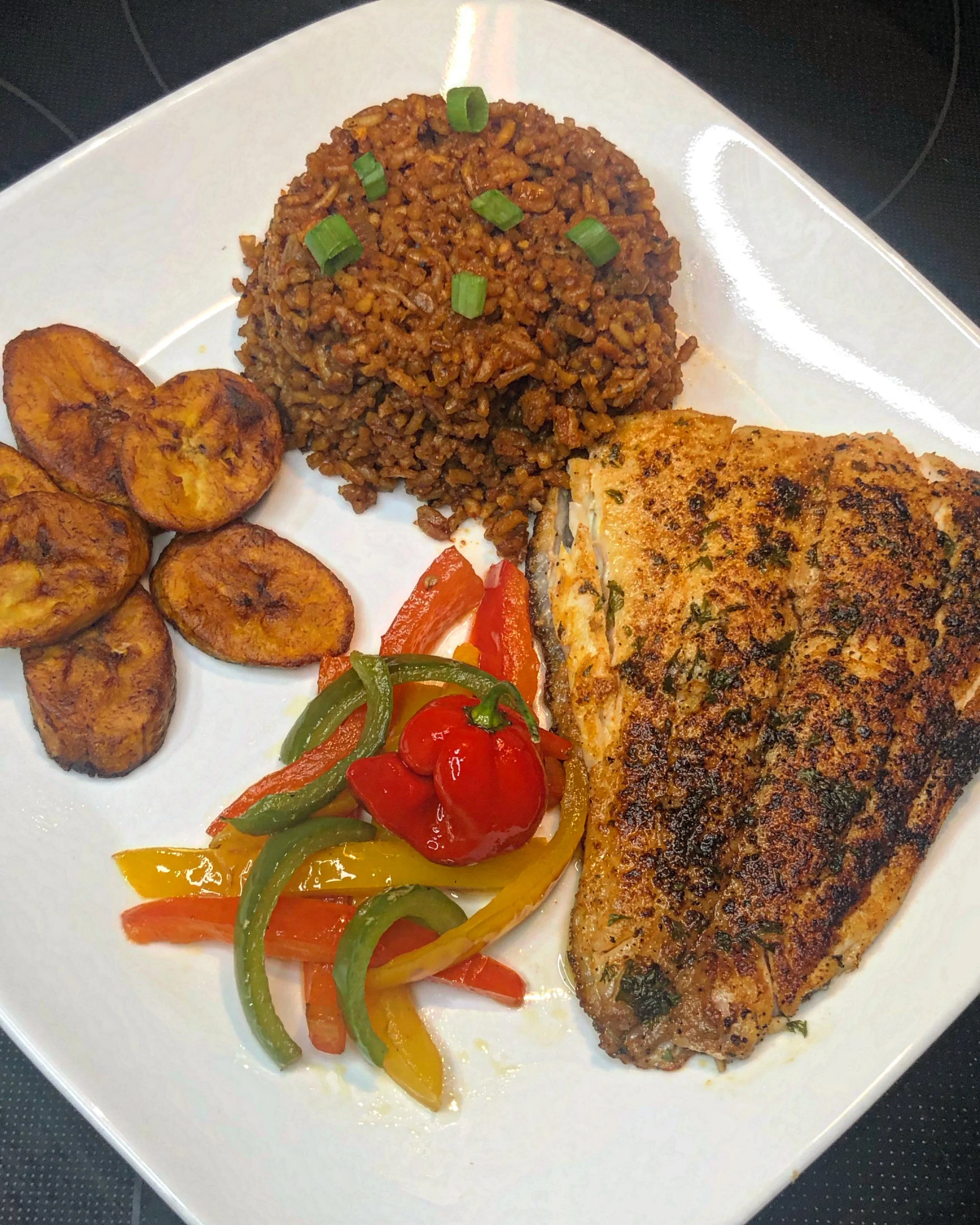 Plate with seasoned grilled fish fillet, seasoned rice topped with green onions, sautéed plantains, and a side of colorful sautéed bell peppers and cherry tomatoes.