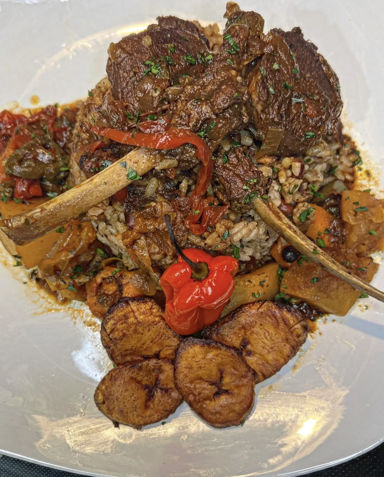 A plate of Jollof rice topped with grilled beef ribs, accompanied by fried plantains, red chili pepper, and vegetables.