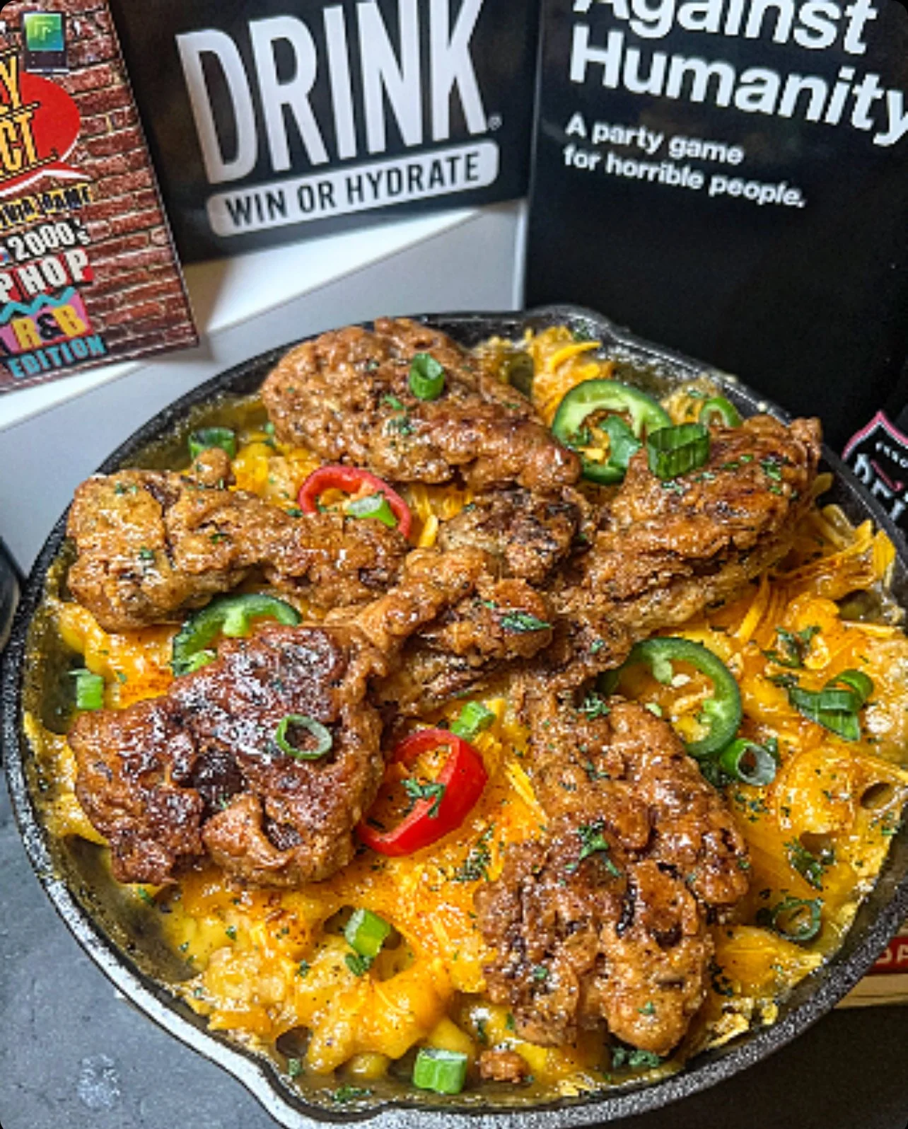 A skillet filled with fried chicken pieces, shredded cheddar cheese, sliced jalapeños, and chopped green onions.