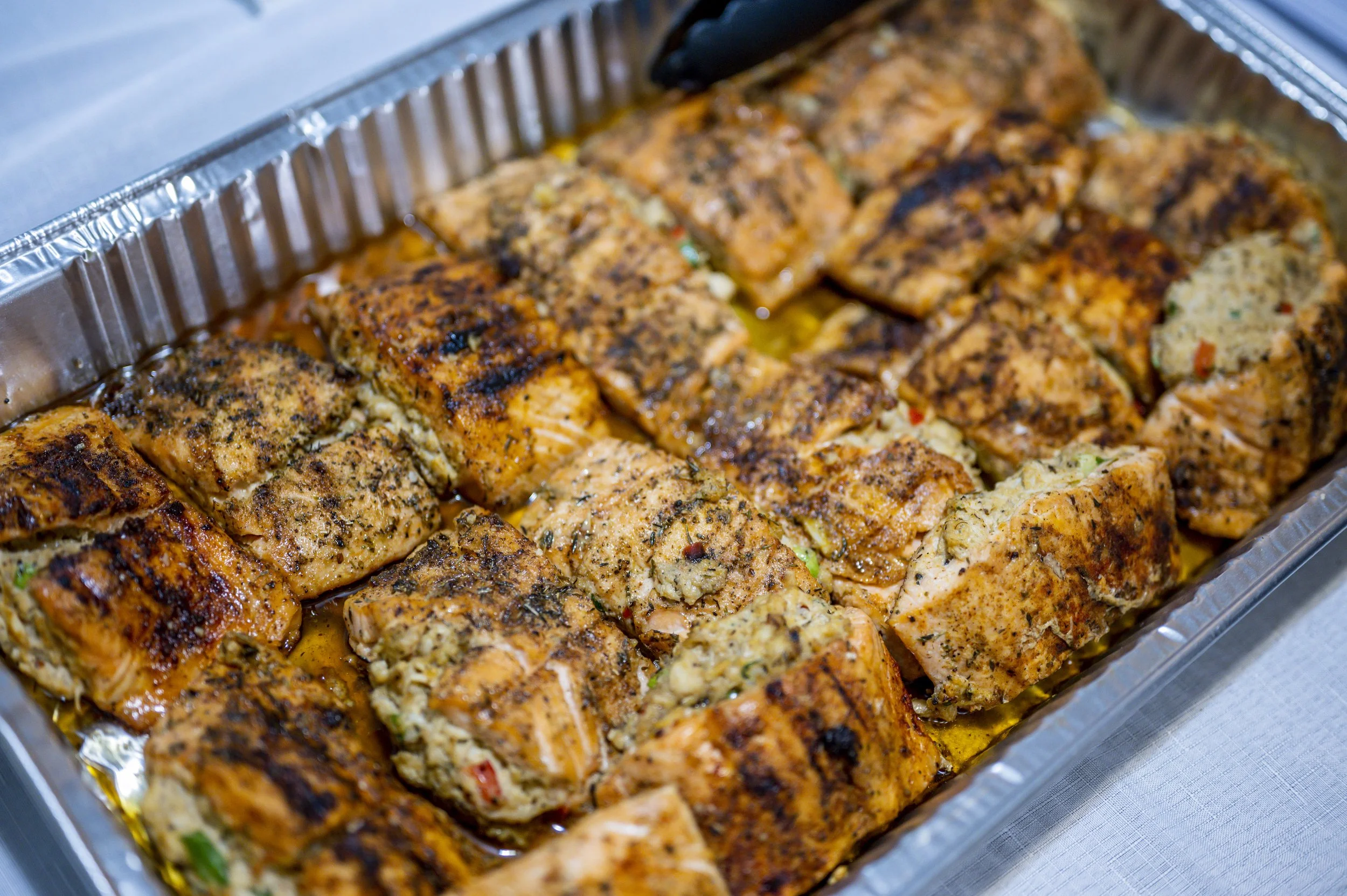 Grilled salmon fillets seasoned and cooked in a metal tray, garnished with herbs.